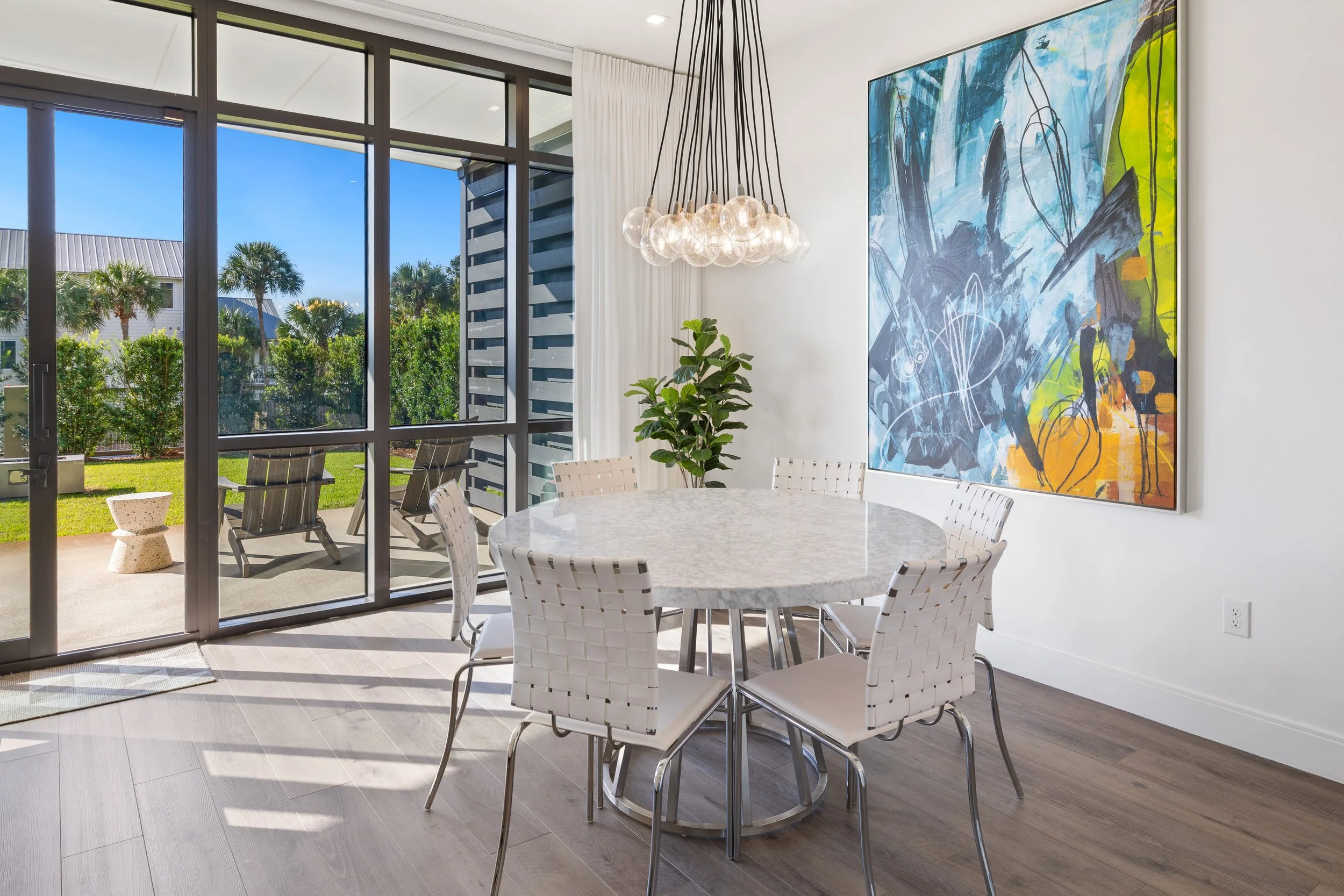 Modern dining room with glass doors opening to outdoor patio with chairs, fenced in yard, and blue sky; round marble table with white woven chairs, colorful abstract artwork on white wall, and a cluster of glass globe pendant lights overhead.