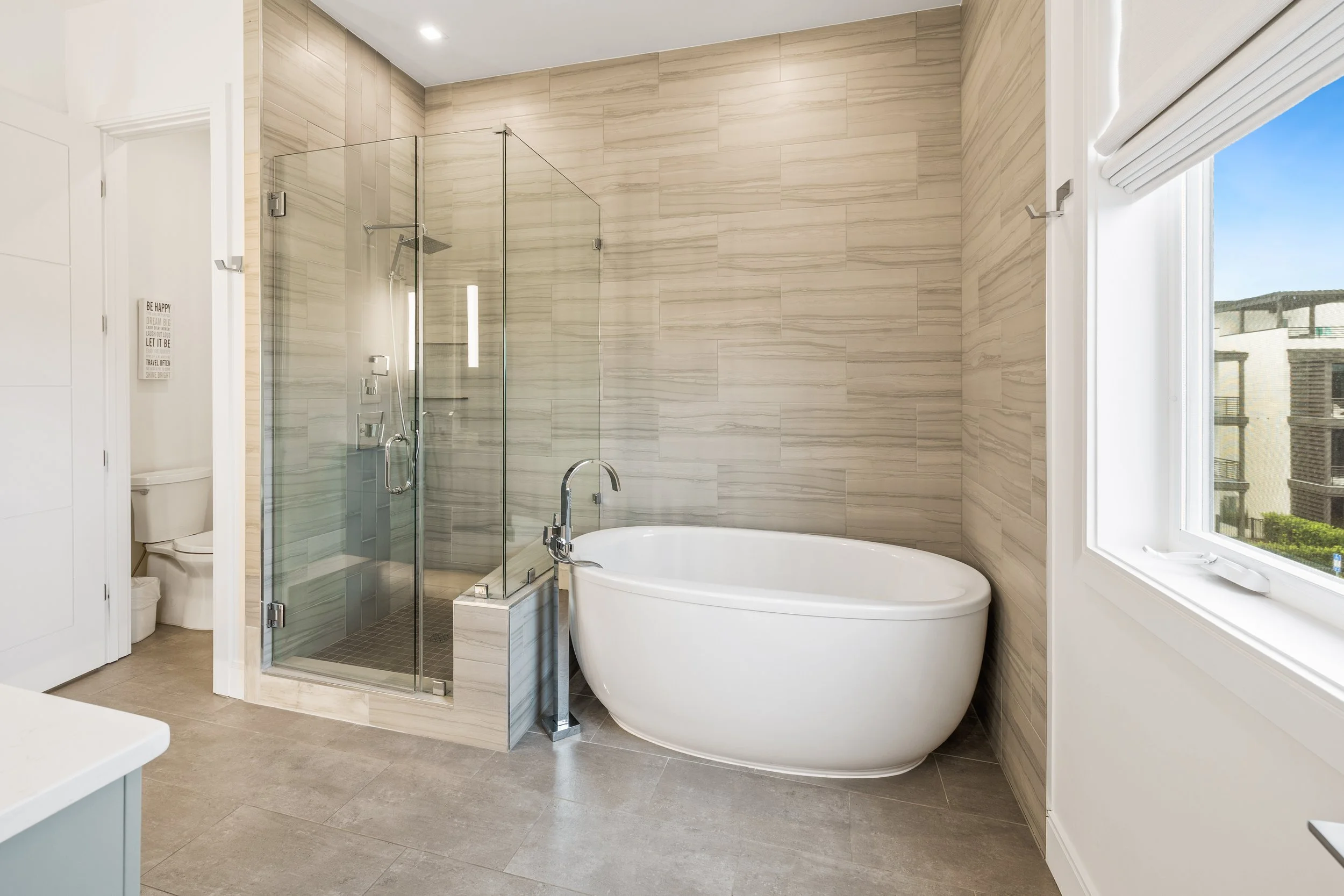 Modern bathroom with a glass-enclosed shower, freestanding bathtub, a window with a view of the pool, and beige tiled walls.