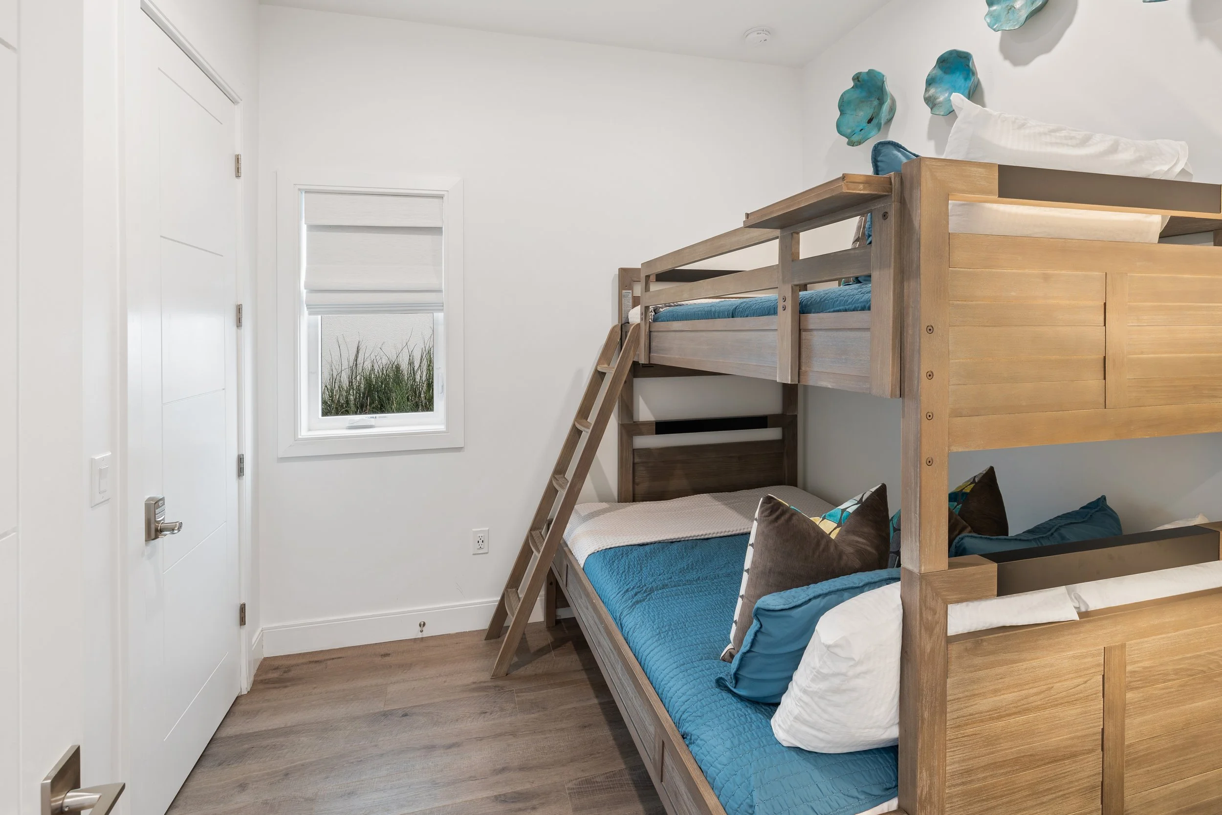 A bedroom with white walls and wooden flooring, featuring a wooden bunk bed with blue bedding and pillows, a small window for natural light.
