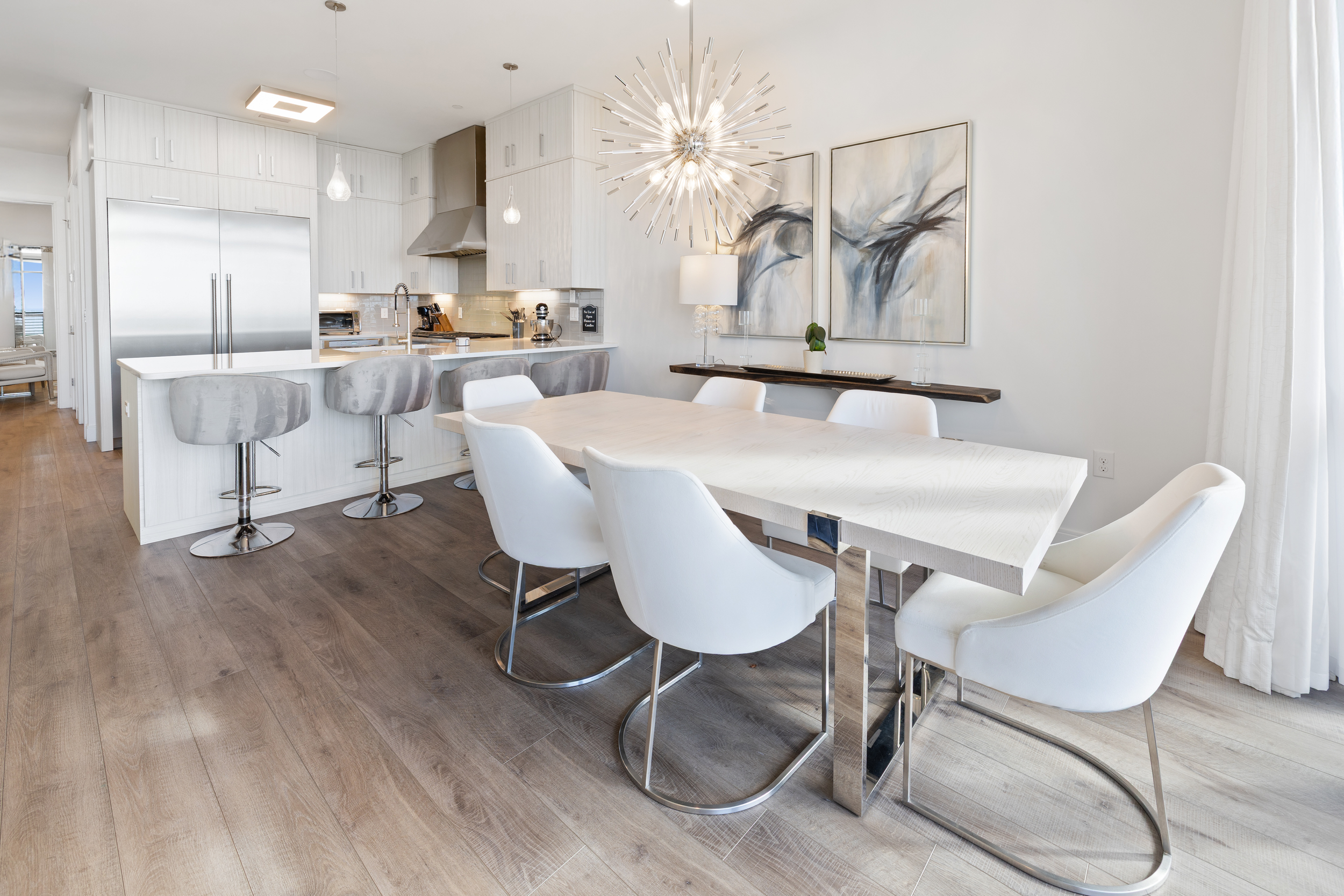 Bright, modern dining area with a white rectangular table, six white chairs, and a large, starburst pendant light. In the background, a kitchen with white cabinetry, stainless steel refrigerator, and a counter with bar stools. 