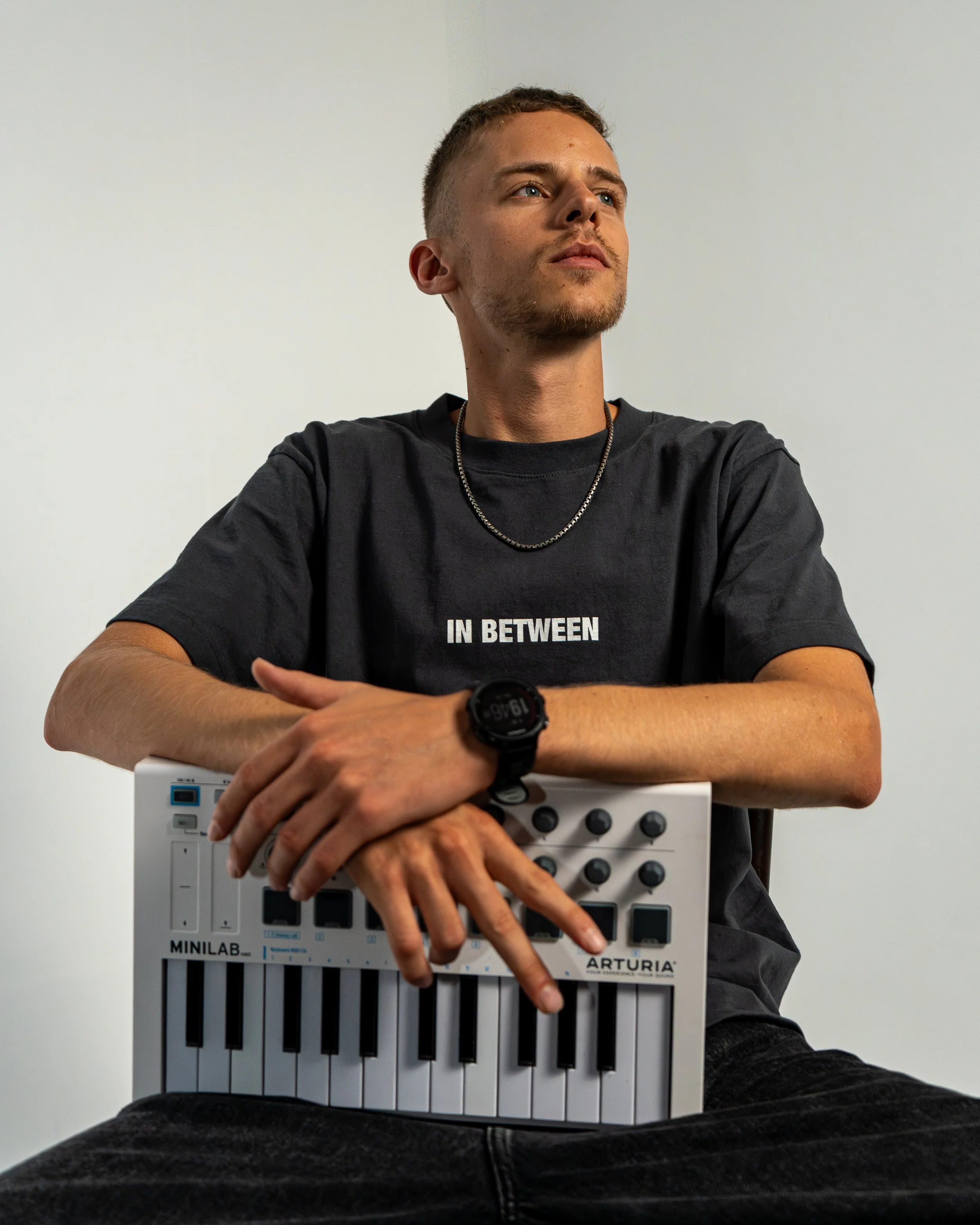 Ein junger Mann sitzt mit einem kleinen MIDI-Controller auf dem Schoß, trägt ein schwarzes T-Shirt mit der Aufschrift 'IN BETWEEN', hat einen silbernen Halskette, eine Armbanduhr und sitzt vor einem neutralen weißen Hintergrund.
