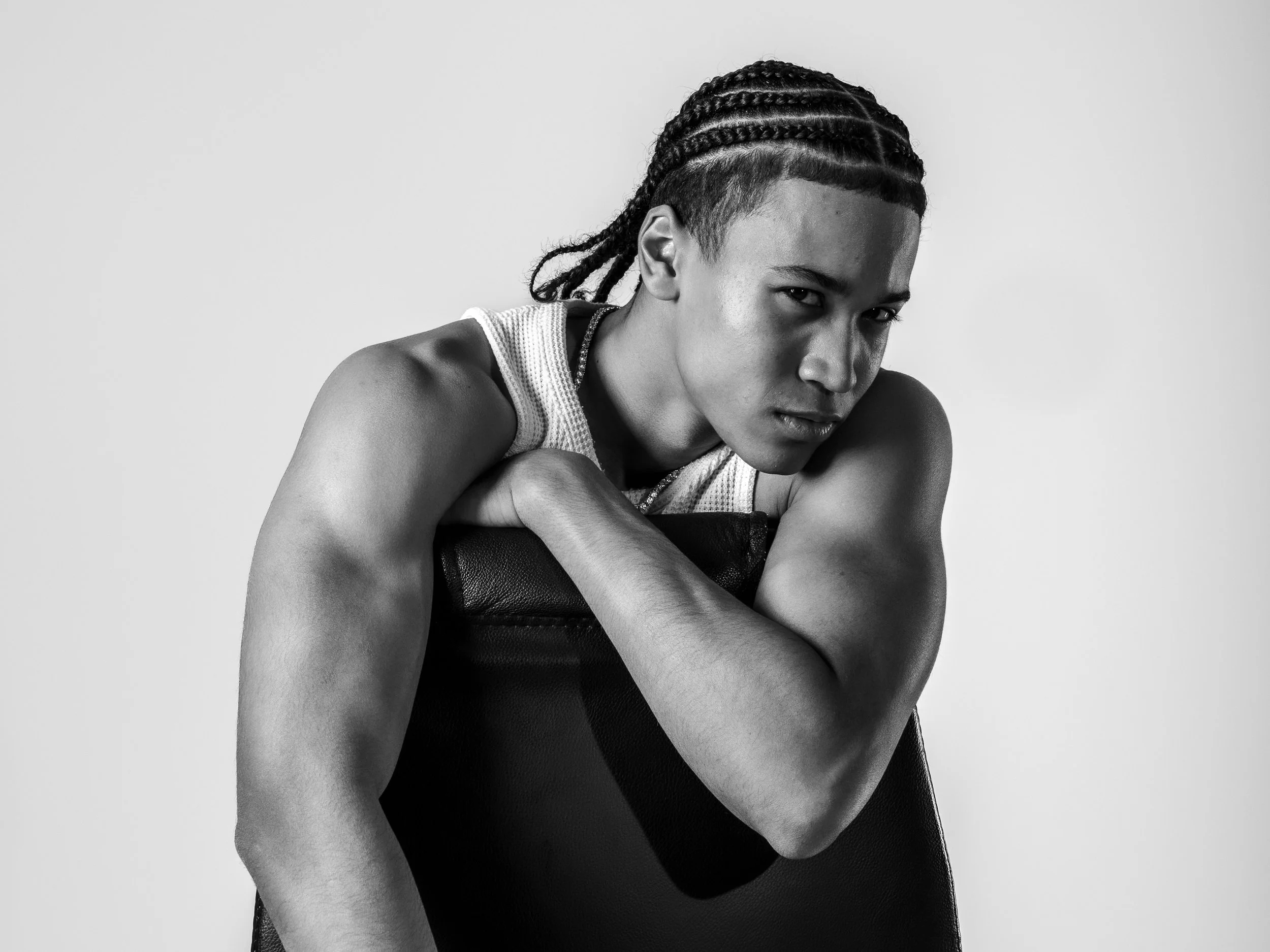 Schwarz-weiß-Fotografie eines jungen Mannes mit dreadlocks, der auf einem schwarzen Stuhl lehnt und direkt in die Kamera schaut.