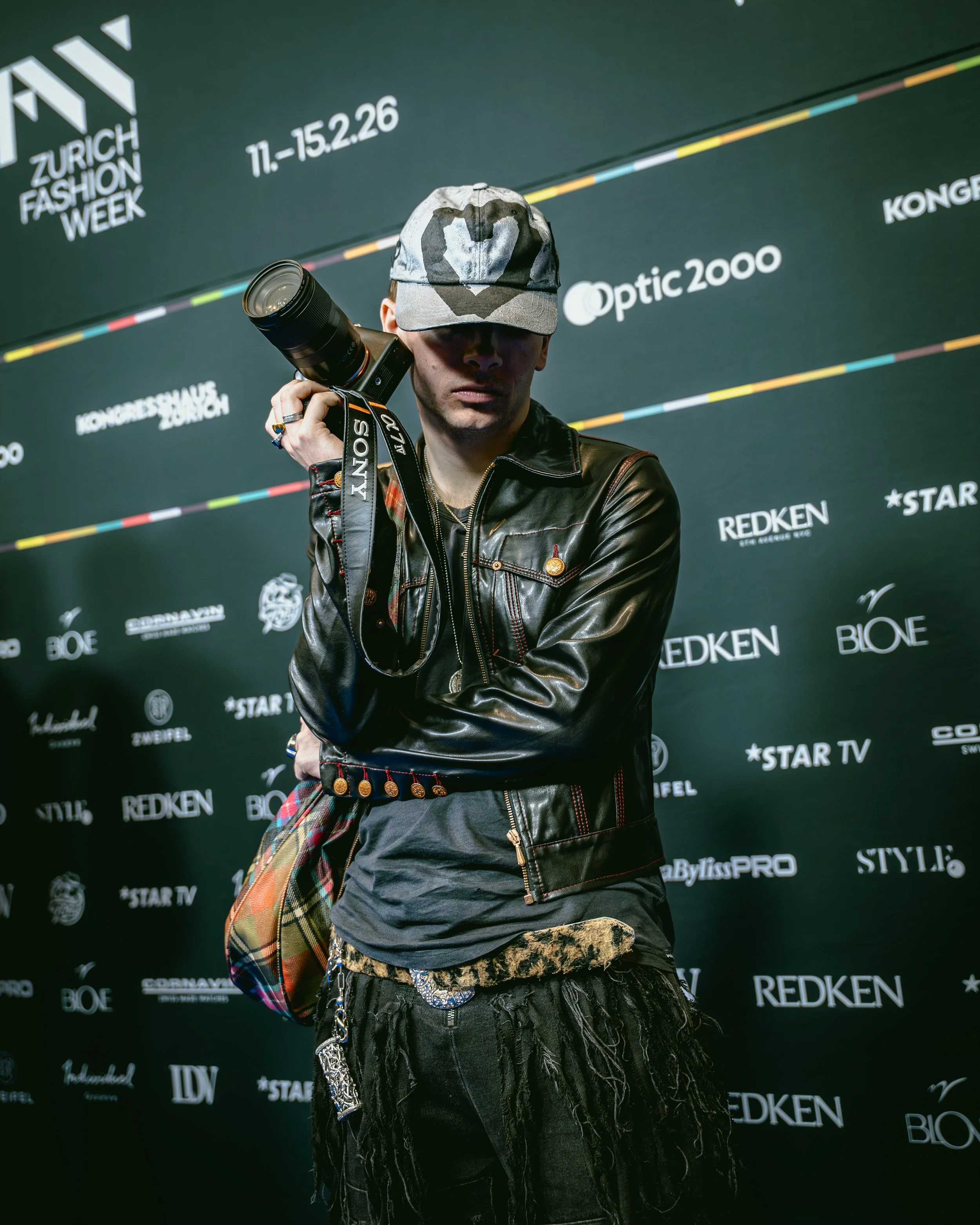 Junge Person mit Camouflage-Mütze und Kamera vor schwarzem Hintergrund mit Logos und Texten, darunter "Zurich Fashion Week".