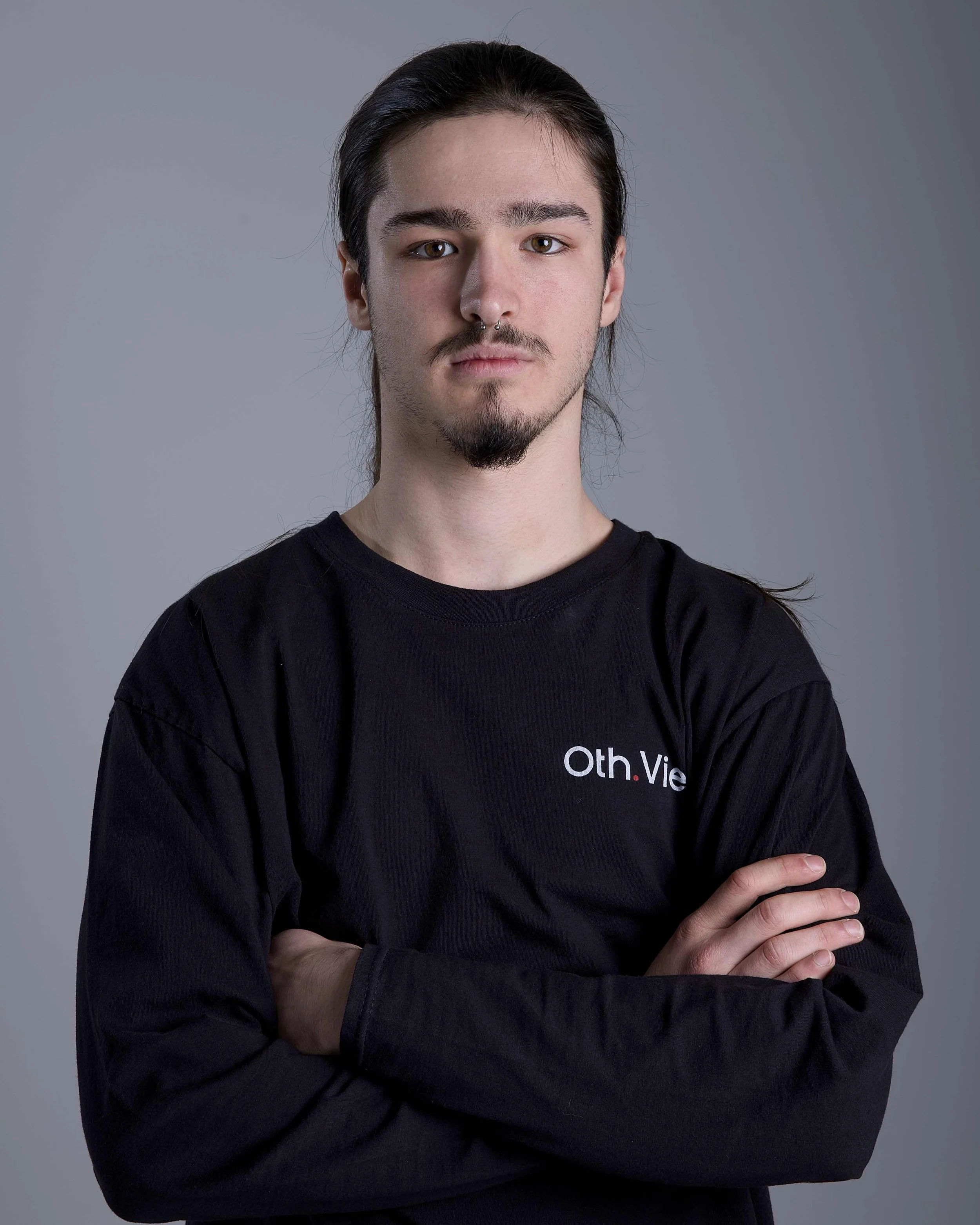 Junge Mann mit langen Haaren, leichtem Bart, im Freien bei bewaldetem Hintergrund.