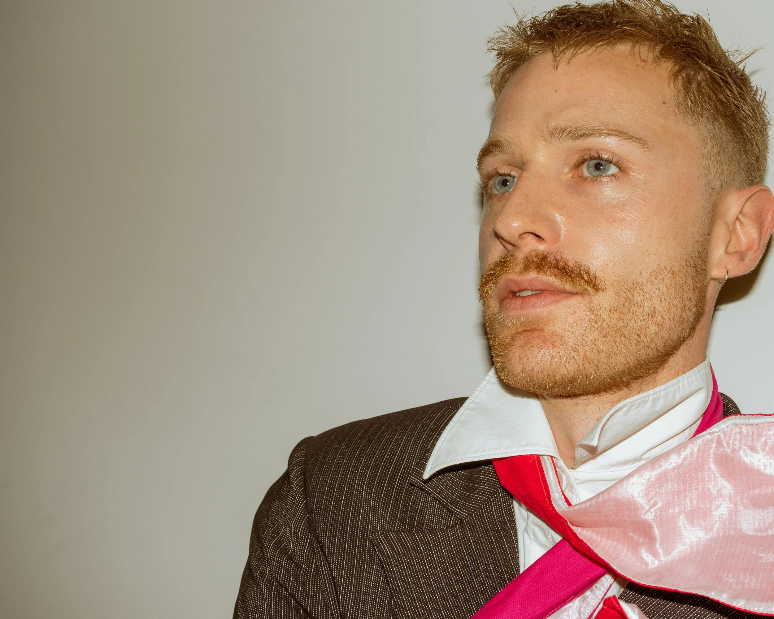 Ein junger Mann trägt ein formelles Hemd und Jacke, mit einem pinken Umhang. Er schaut nach rechts, vor einem neutralen Hintergrund.