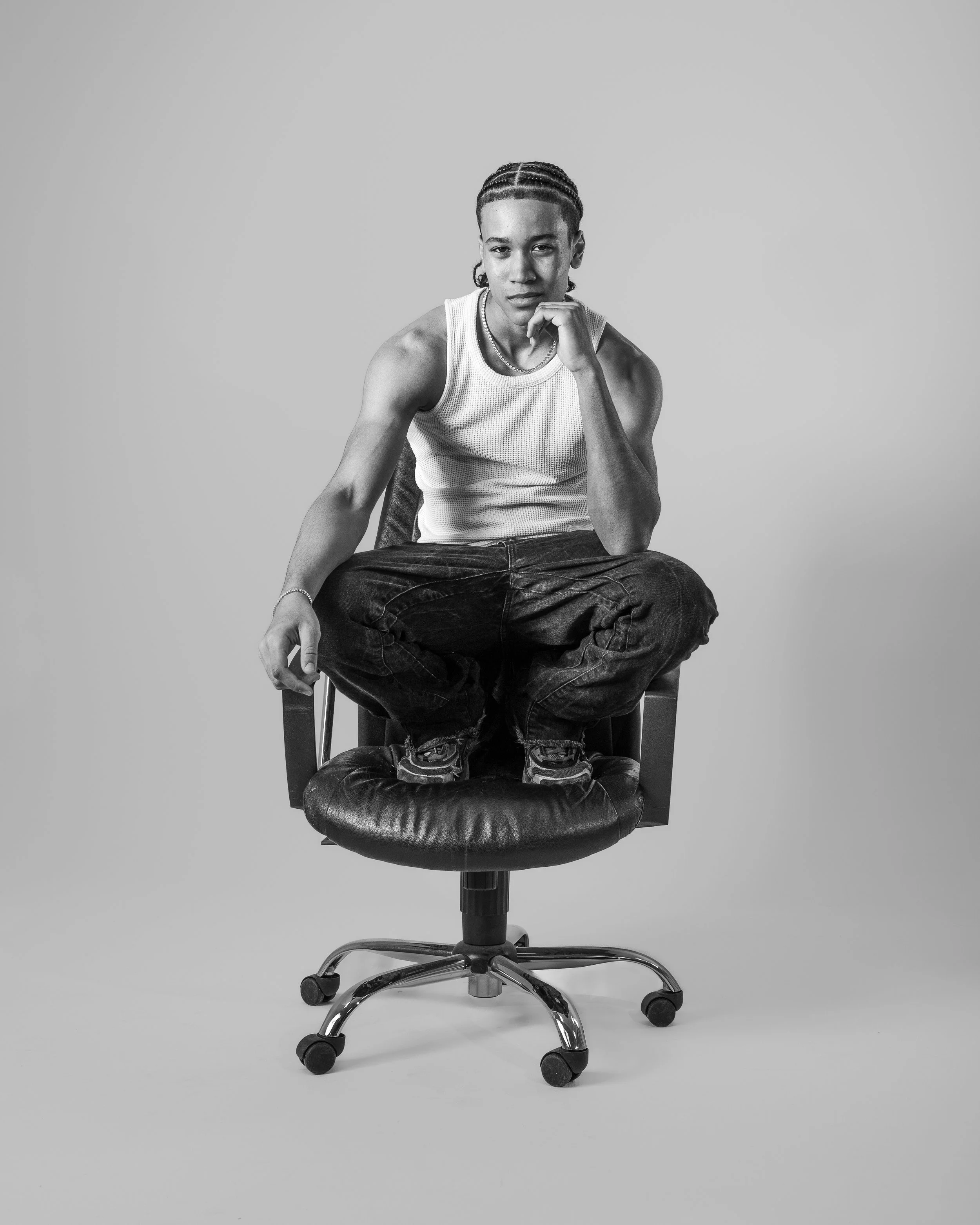 Schwarzer und weißer Portrait eines jungen Mannes, der auf einem Bürostuhl sitzt, mit einem nachdenklichen Ausdruck, Arme verschränkt, kurze geflochtene Haare, weiße ärmellose T-Shirt, dunkle Hosen, auf einem neutralen Hintergrund.