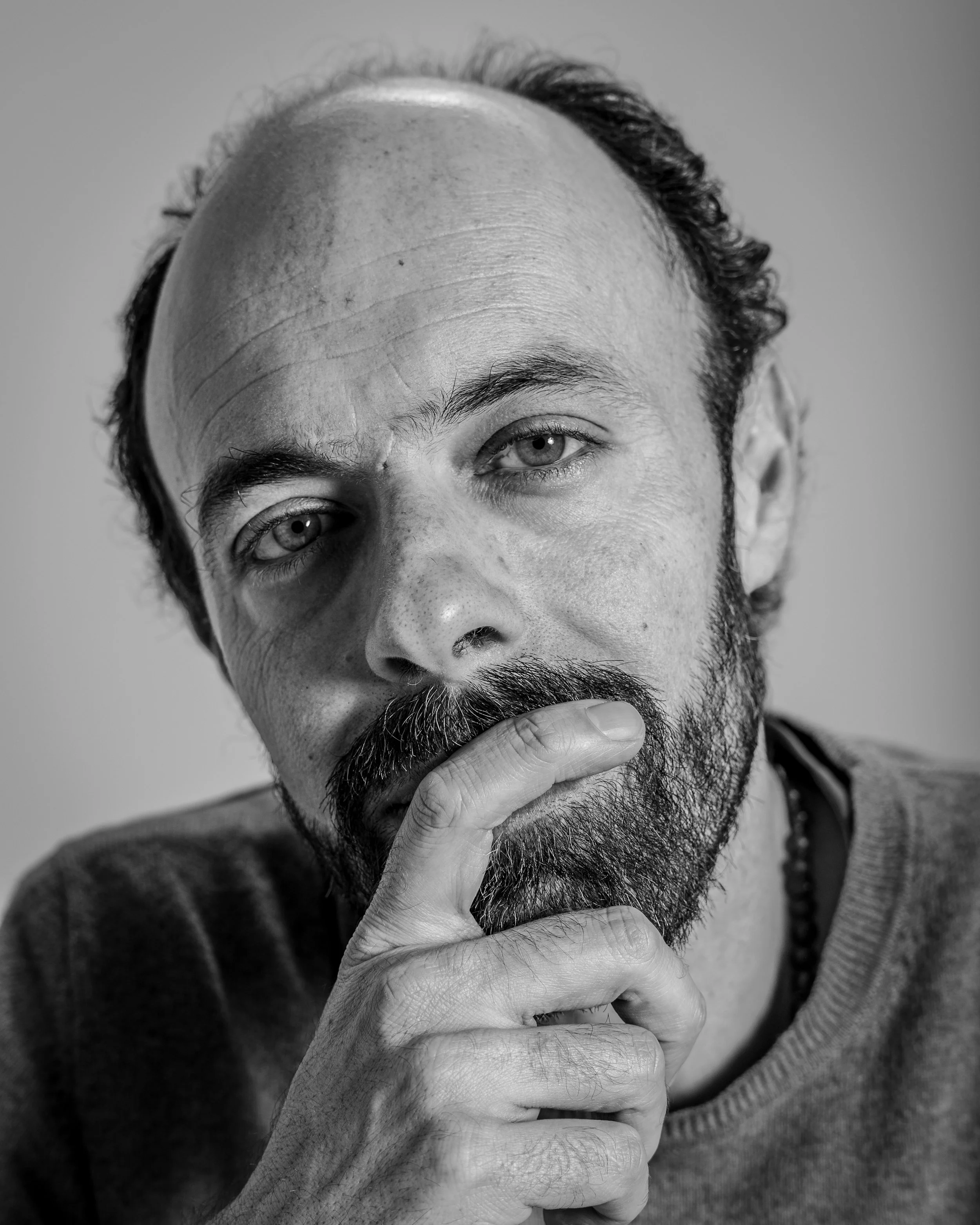 Schwarz-Weiß-Fotografie eines Mannes mit Halbglatze, Bart und nachdenklicher Pose, die Hand an den Mund gelegt.