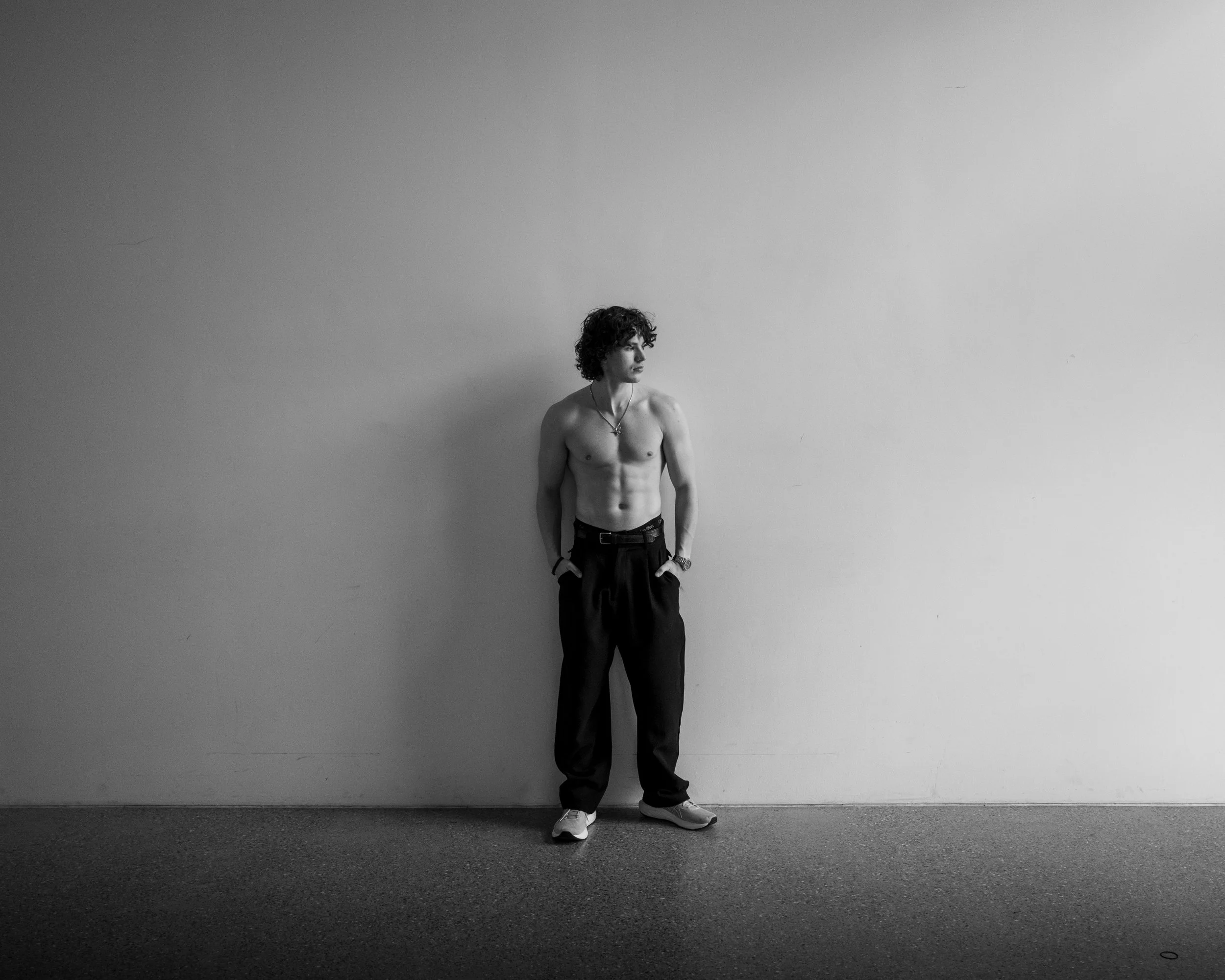 Schwarz-weisses Foto eines jungen Mannes, der nur eine Halskette trägt, in schwarzen Hosen und Turnschuhen, an einer leeren weißen Wand lehnt, mit kurzen, lockigen Haaren und einem nachdenklichen Blick nach links gerichtet.