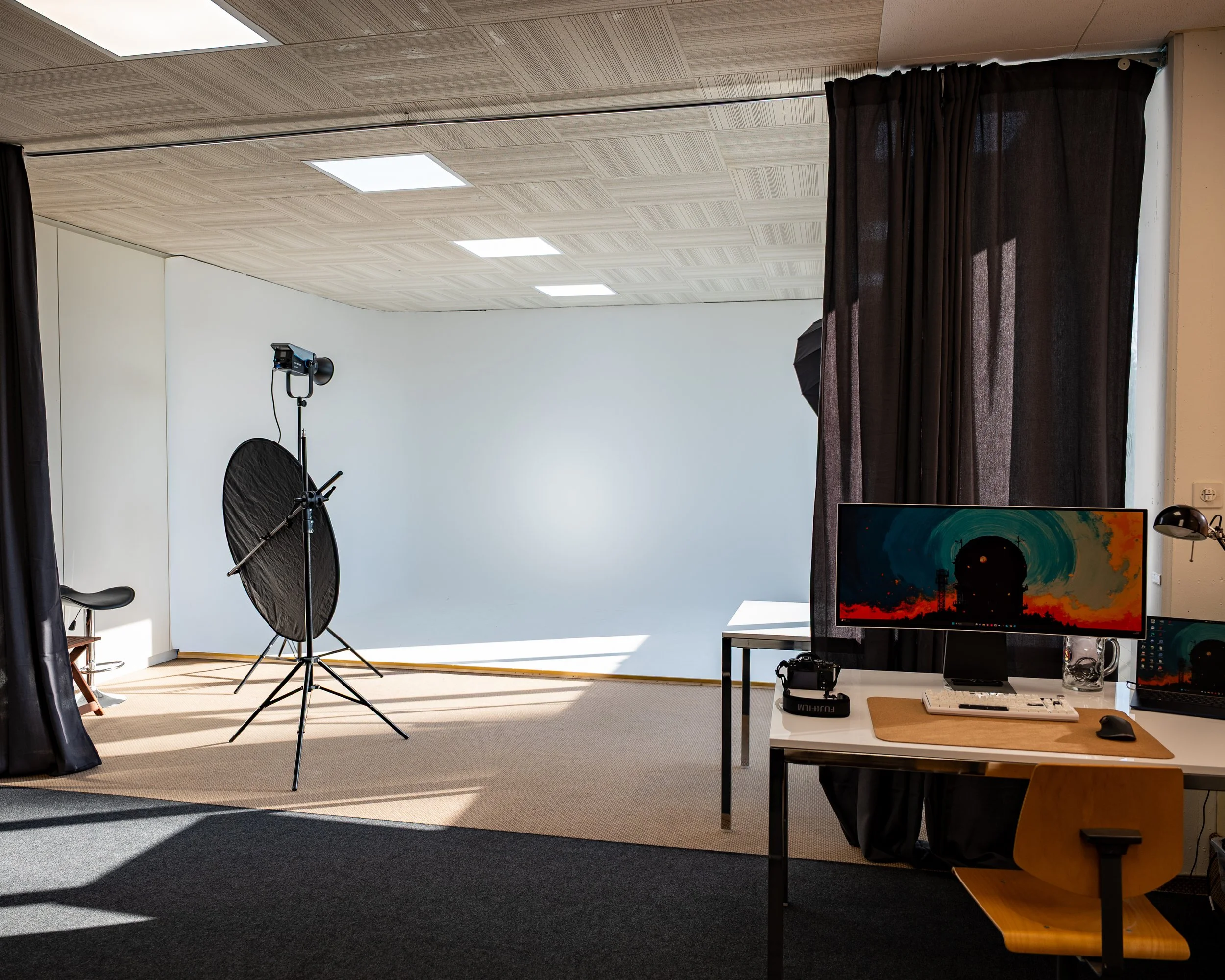 Fotostudio mit professioneller Beleuchtung, schwarzem Hintergrund, Computern, Monitor und Sitzgelegenheit im Raum.