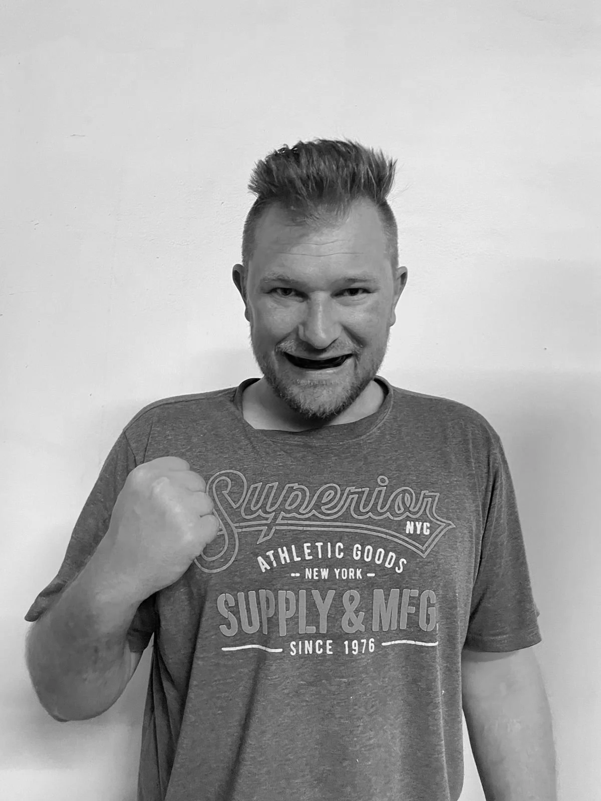 A man with short spiked hair and a beard, smiling and making a fist gesture, wearing a graphic T-shirt, in front of a plain wall.