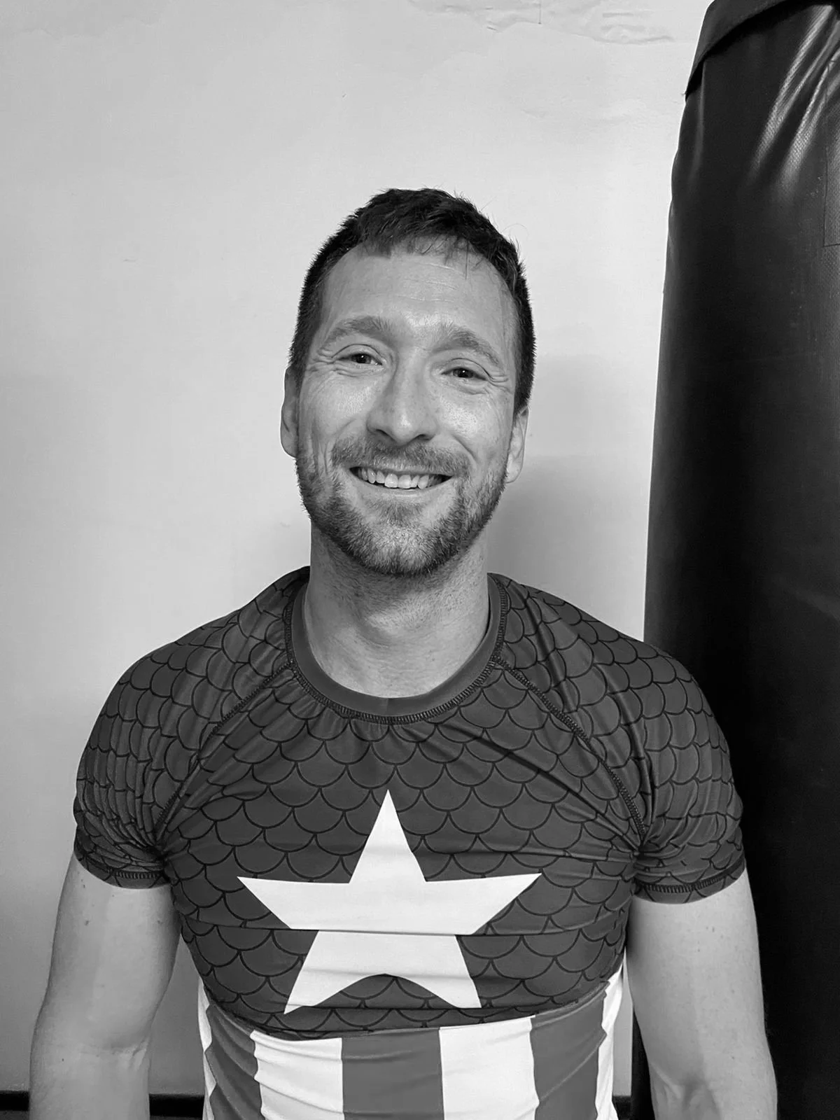 A smiling man with short dark hair and facial hair, wearing a sports shirt with a star and striped pattern.