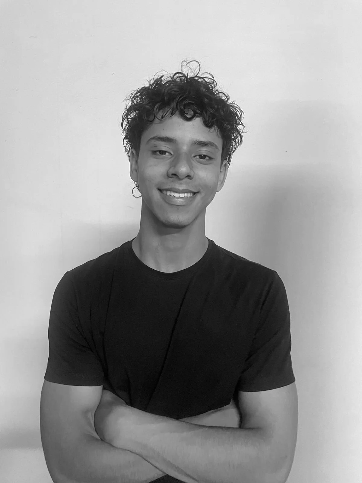A young man with curly hair smiling, wearing a black t-shirt, arms crossed, standing against a plain background.