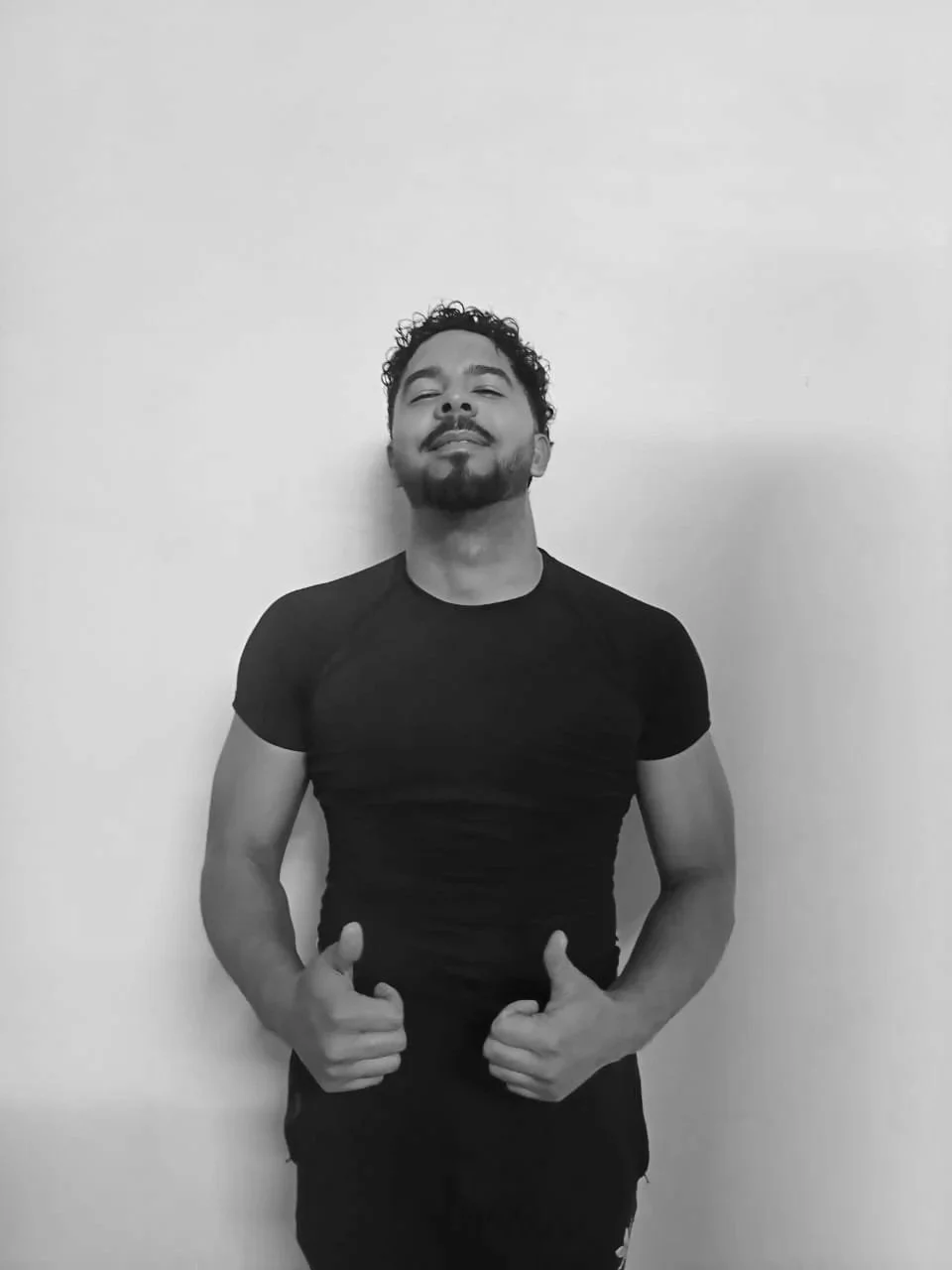A man with curly hair and a beard giving a thumbs-up with both hands, wearing a black t-shirt, standing against a plain wall.