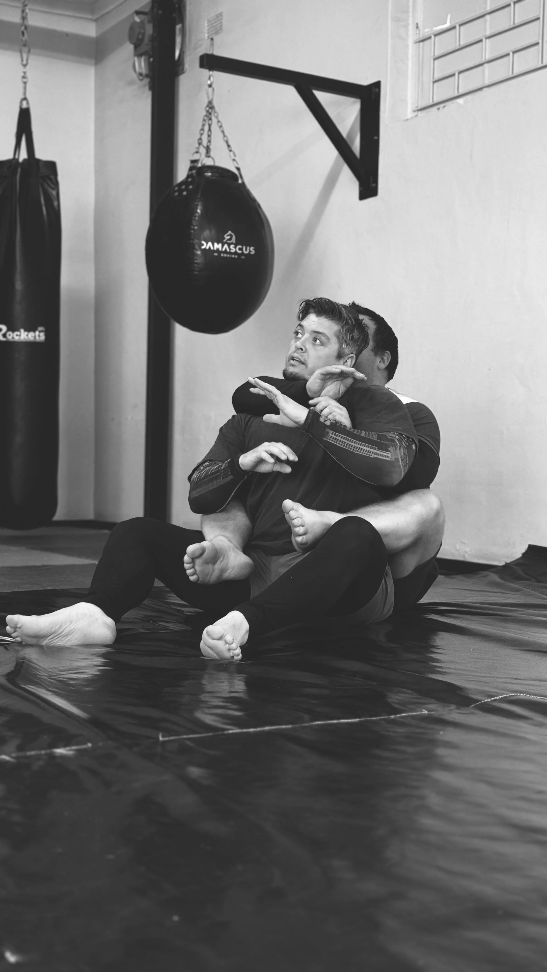 Two men practicing Brazilian Jiu-Jitsu on a black mat in a gym, with one man applying a chokehold on the other.
