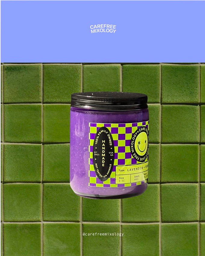 A purple lavender-scented candle in a jar with a black lid, attached to a green tiled wall. The label has yellow and purple checks and says 'Handmade' and 'Lavender.' At the top, there is a blue sign with white text that reads 'Carefree Mixology.'
