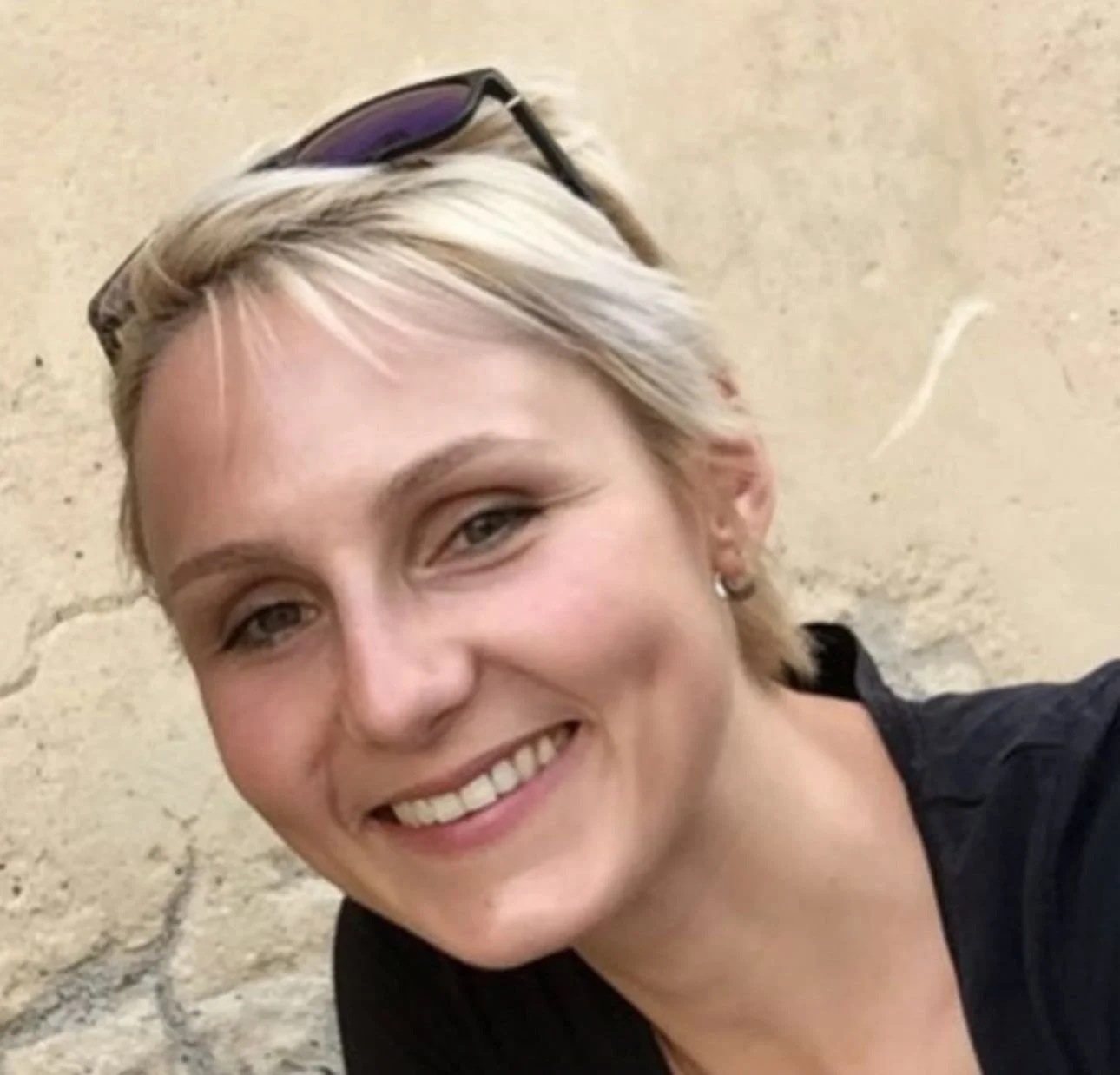Close-up of a smiling woman with short blonde hair, sunglasses on her head, wearing earrings and a dark top, in front of a beige brick wall.