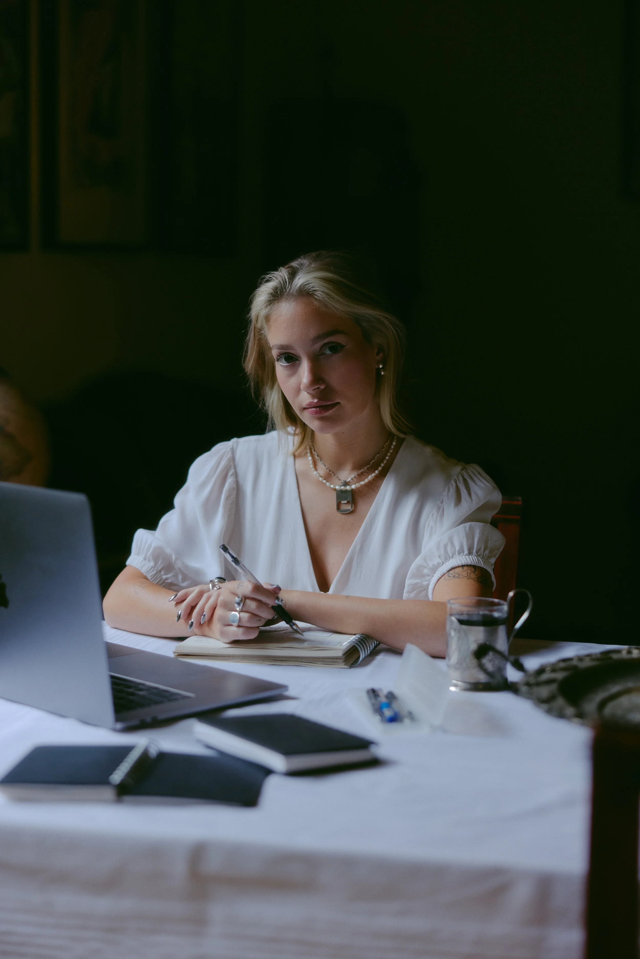 Woman with blonde hair and jewelry sitting at a desk with a laptop, notebooks, papers, and a coffee mug.