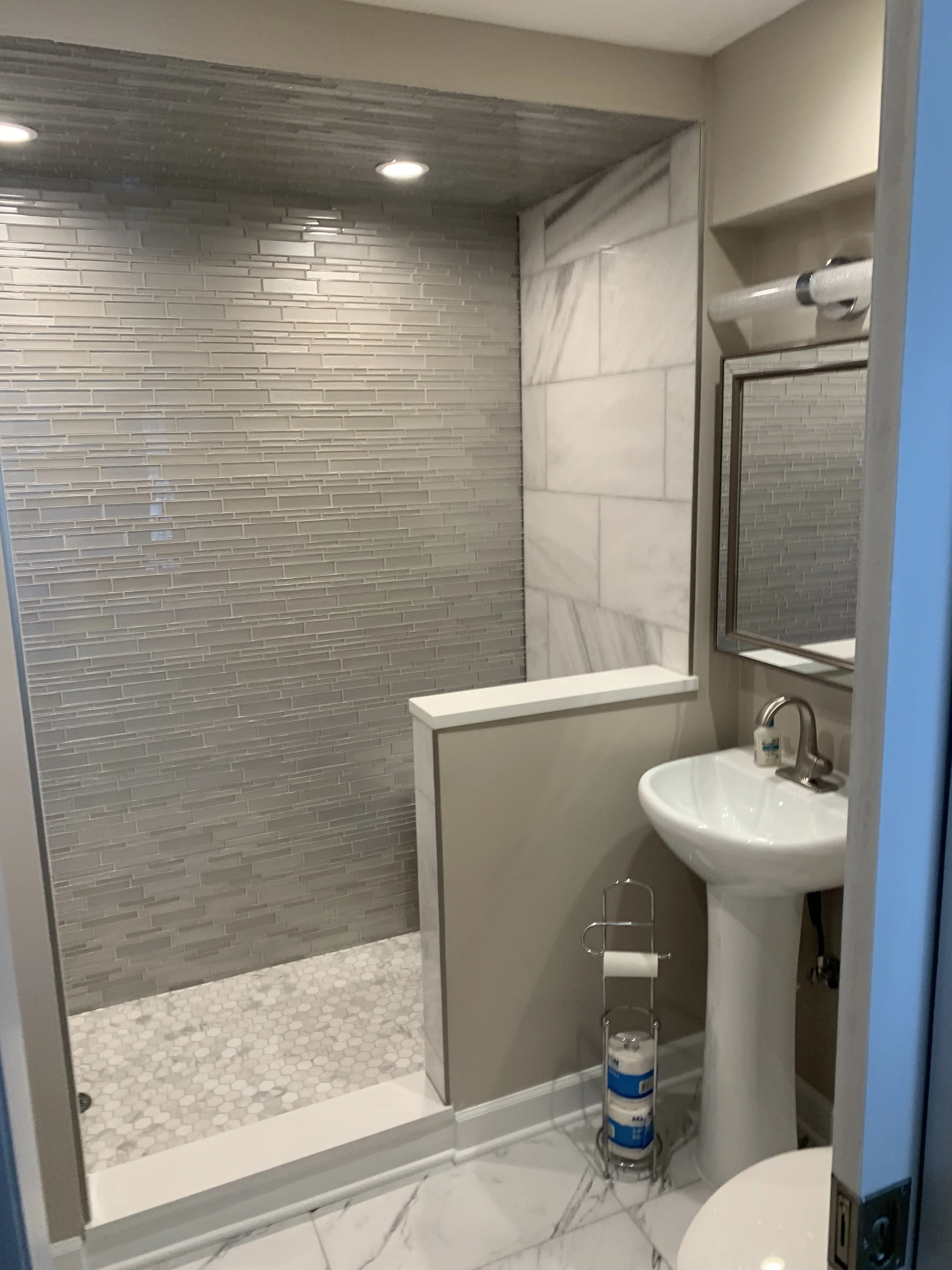 A small bathroom with a walk-in shower, a white pedestal sink with a mirror above, and a toilet with a toilet paper holder near it. The shower has a grey textured tile wall and a small hexagon tile floor, separated by a partial wall.