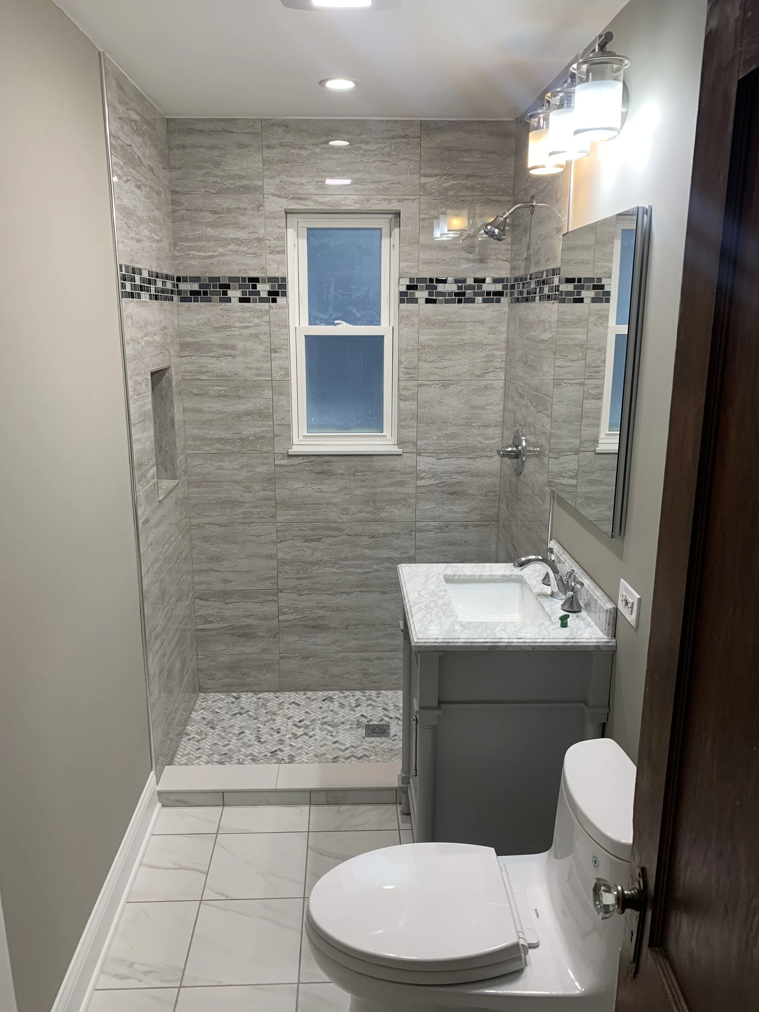 Small bathroom with a walk-in shower, white toilet, gray vanity with marble countertop, mirror, frosted window, and white tile flooring.