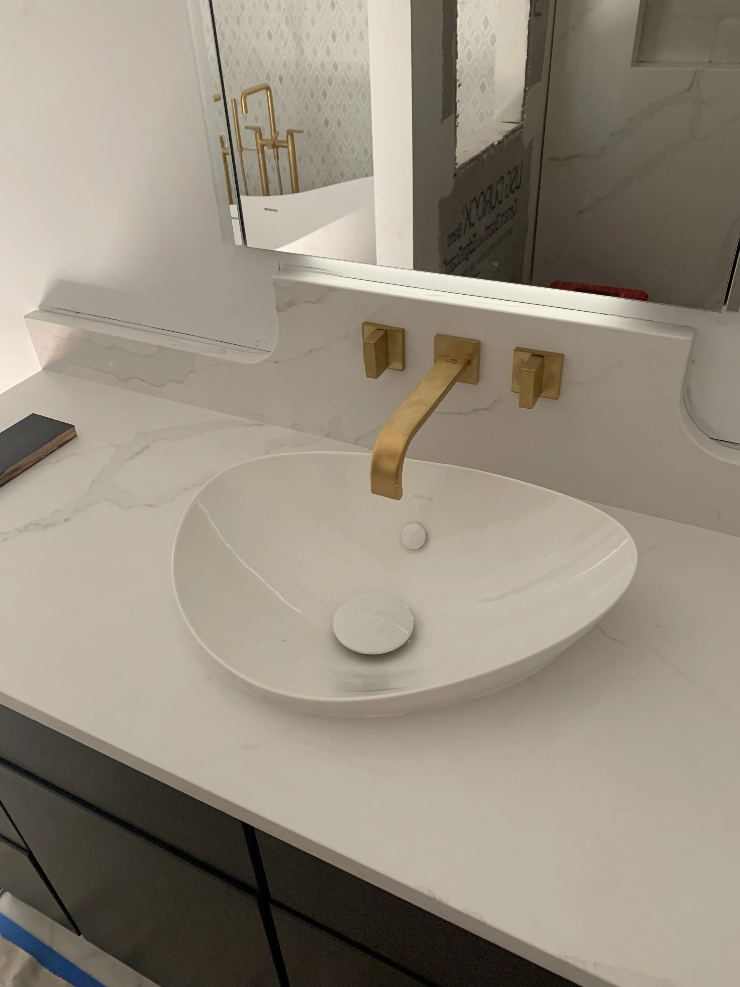 A modern bathroom sink with a white marble countertop, a white oval vessel sink, and a gold faucet with matching gold handles. There is a mirror above the sink and a black book to the left on the countertop.