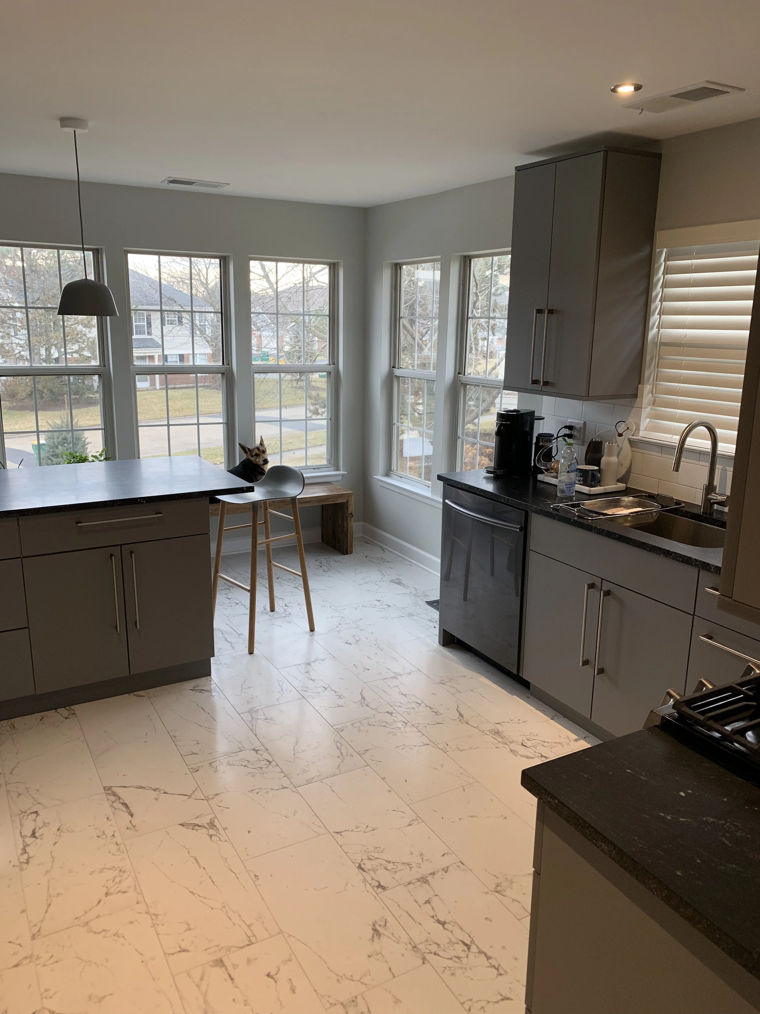 Kitchen with gray cabinets, black countertop, marble-style tile floor, a window nook with a small table, and a cat sitting on a stool near the window.