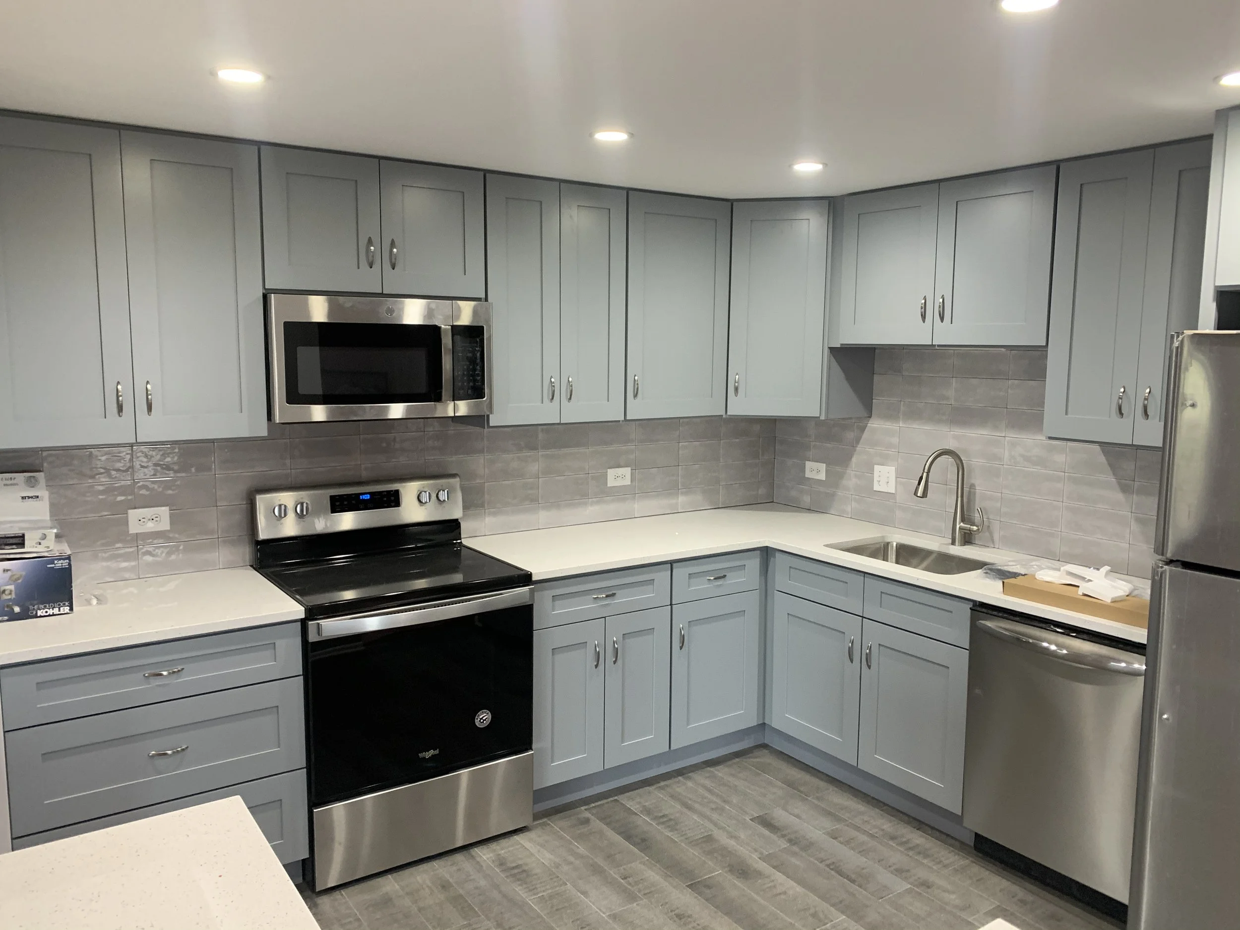 Modern kitchen with light blue cabinets, stainless steel appliances, gray tile backsplash, and gray wood-look flooring.