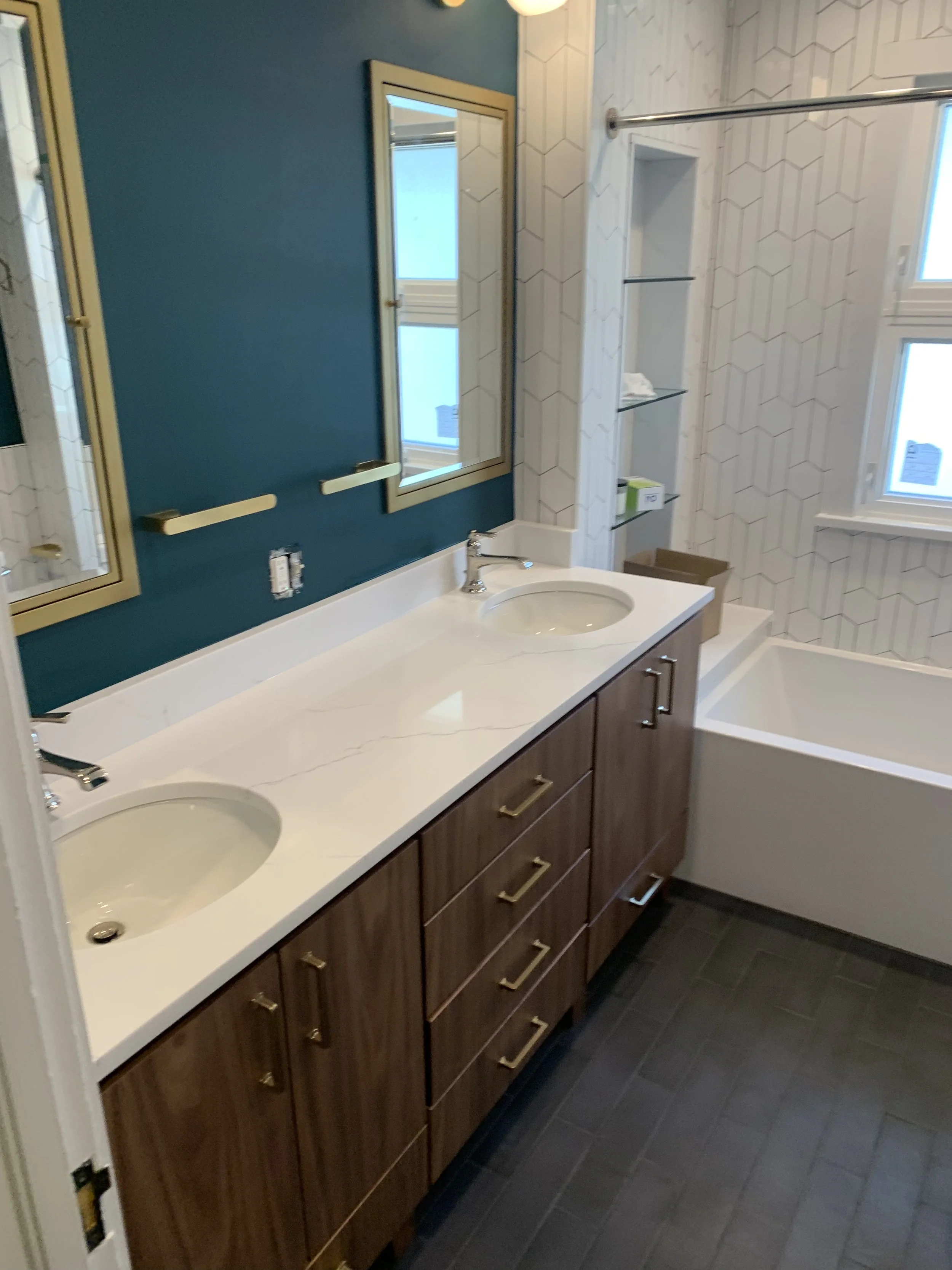 Modern bathroom with a dual-sink vanity, teal accent wall with gold-framed mirrors, shelving, a bathtub, and hash patterned white tiles.