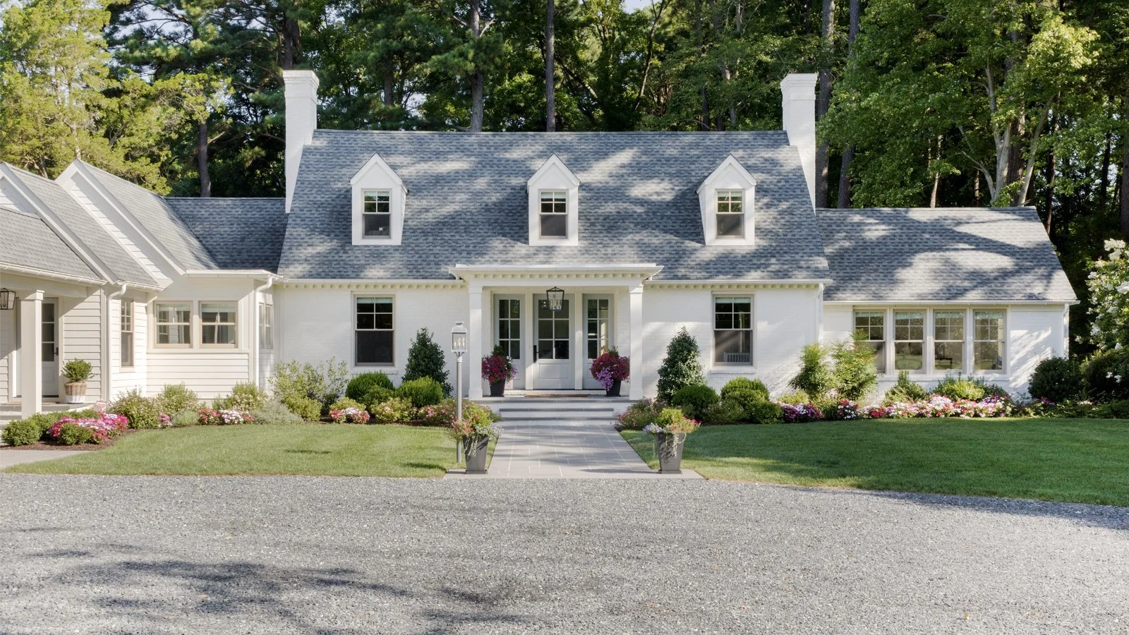 Front view of a white house with a gray shingle roof, surrounded by a lush green lawn and garden with pink and purple flowers, and large trees in the background.