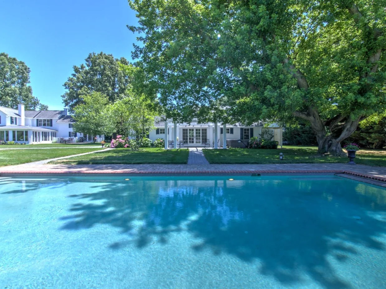 View of a backyard with a swimming pool in the foreground, a large tree, and a white house with a porch in the background under a clear blue sky.
