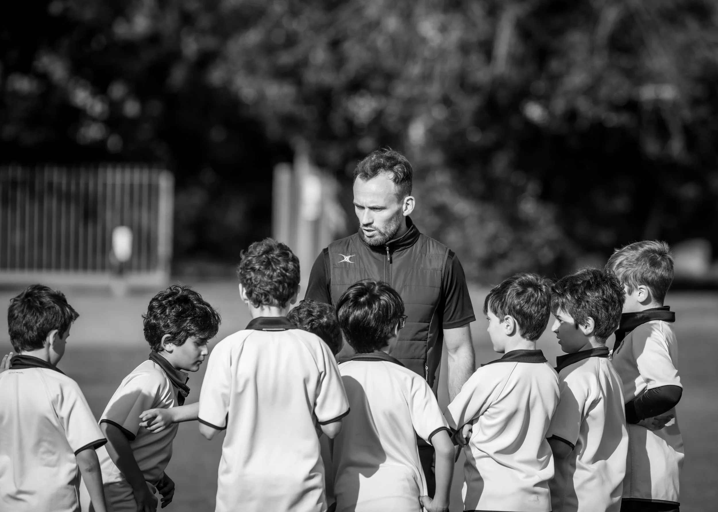 A coach giving instructions to a group of young soccer players huddled together on a field during daytime, black and white photo.