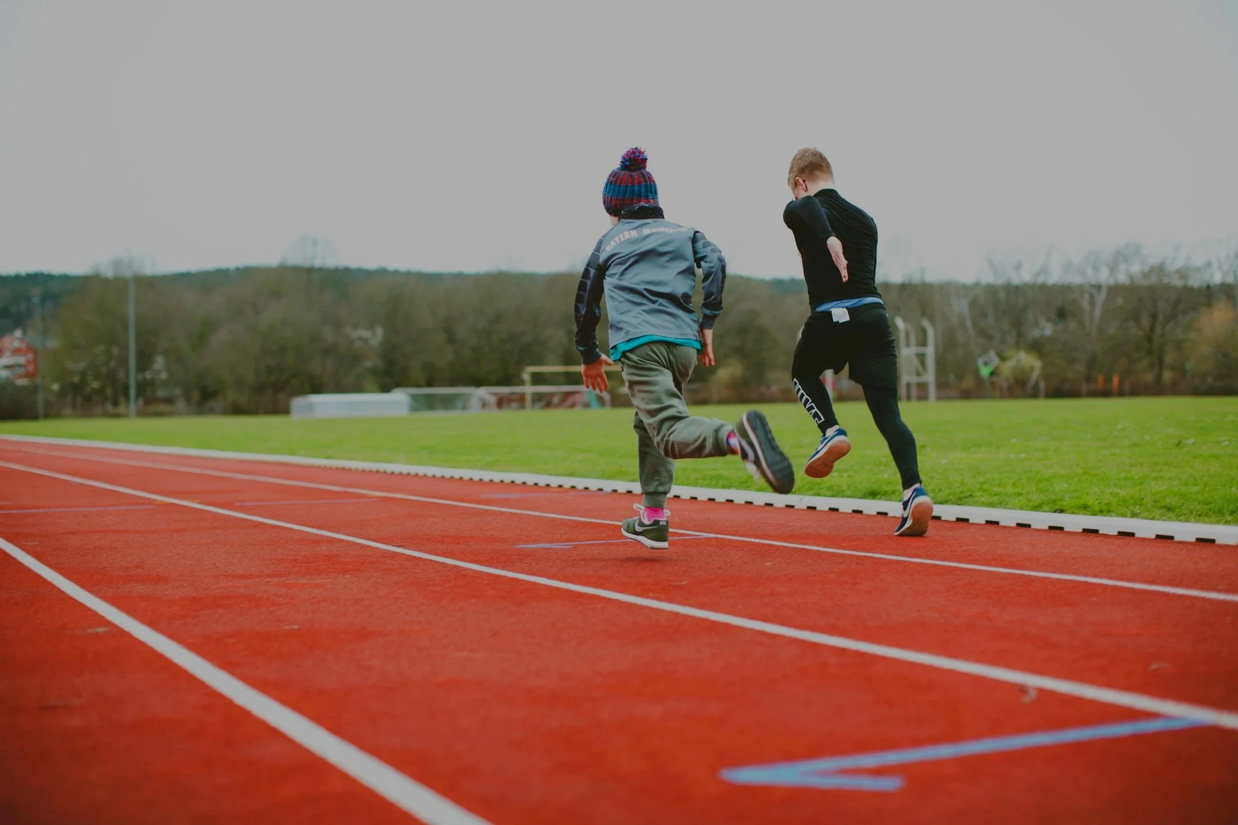 Two children running on a red track at a sports field, with green grass and trees in the background.