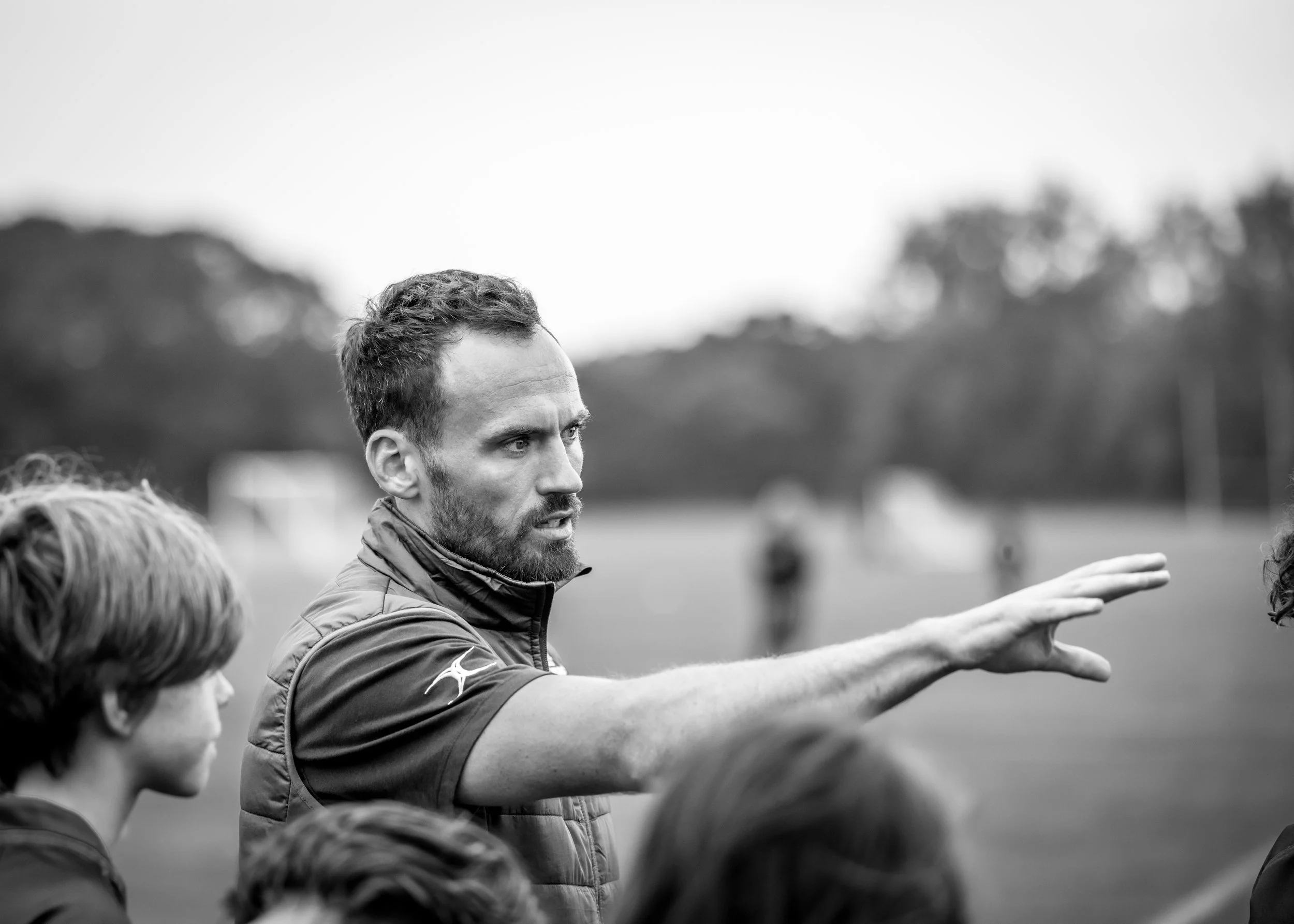 A black and white photograph of a man with short hair and beard, wearing a sports jacket, gesturing with his right arm extended, speaking to a group of people outdoors on a field, with trees and tents in the background.