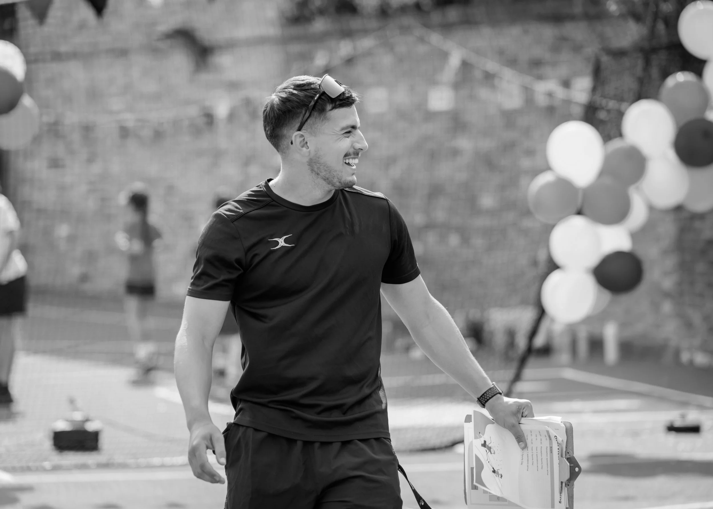A young man in athletic clothing smiling and holding a clipboard, standing outdoors with balloons in the background.