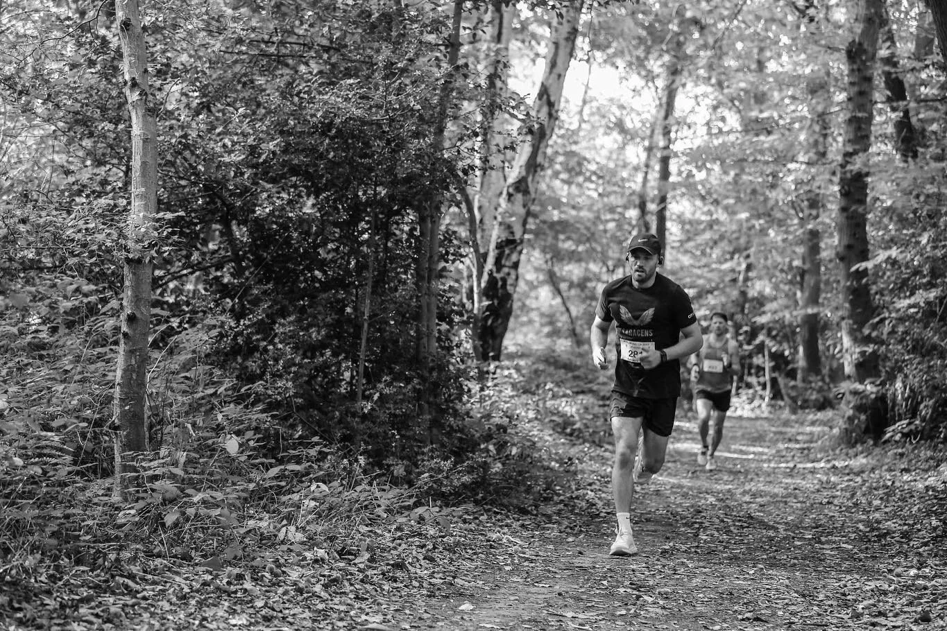 Two men running on a trail through a forest during a race, with trees and foliage surrounding them.