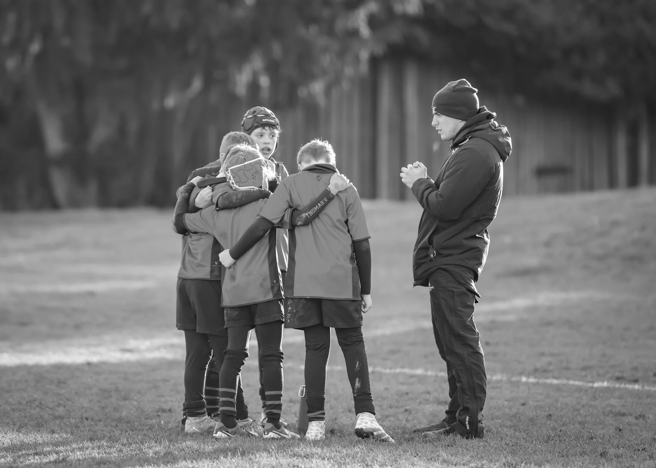 A group of young boys, dressed in sports attire, gathered in a huddle on a grassy field during daytime. An adult coach stands nearby, with his hands clasped, addressing them.