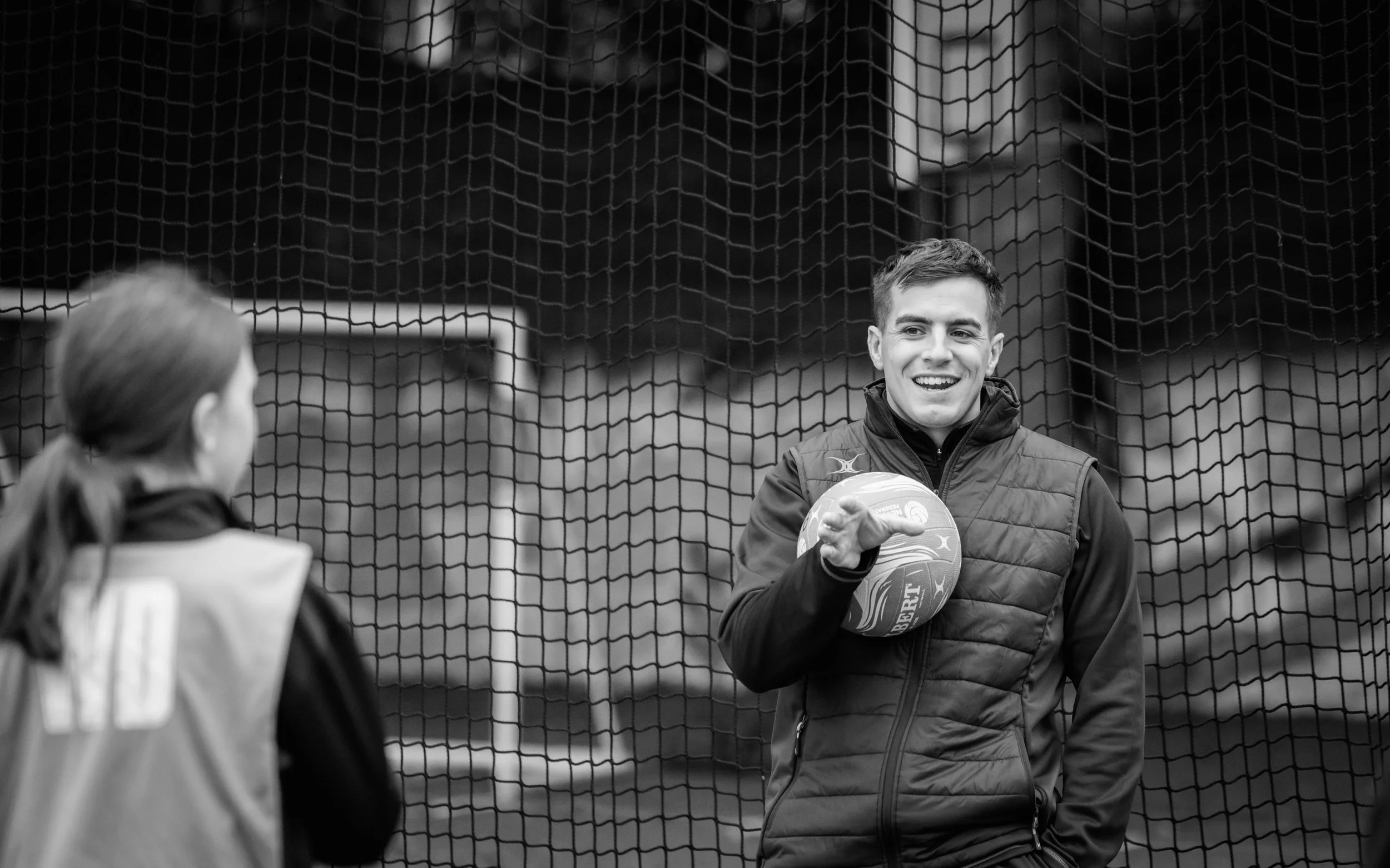 A young man holding a basketball and smiling while talking to a woman inside a sports net on a sports field.