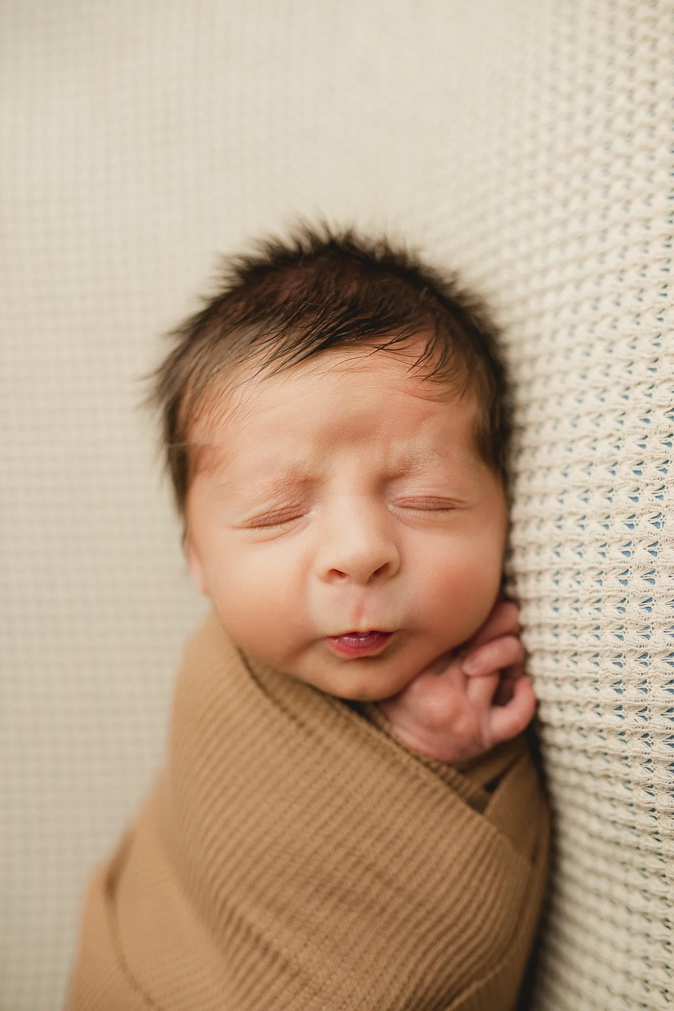 Close-up of a sleeping baby's face with eyes closed, resting against a textured surface, with hands gently clasped near the face.