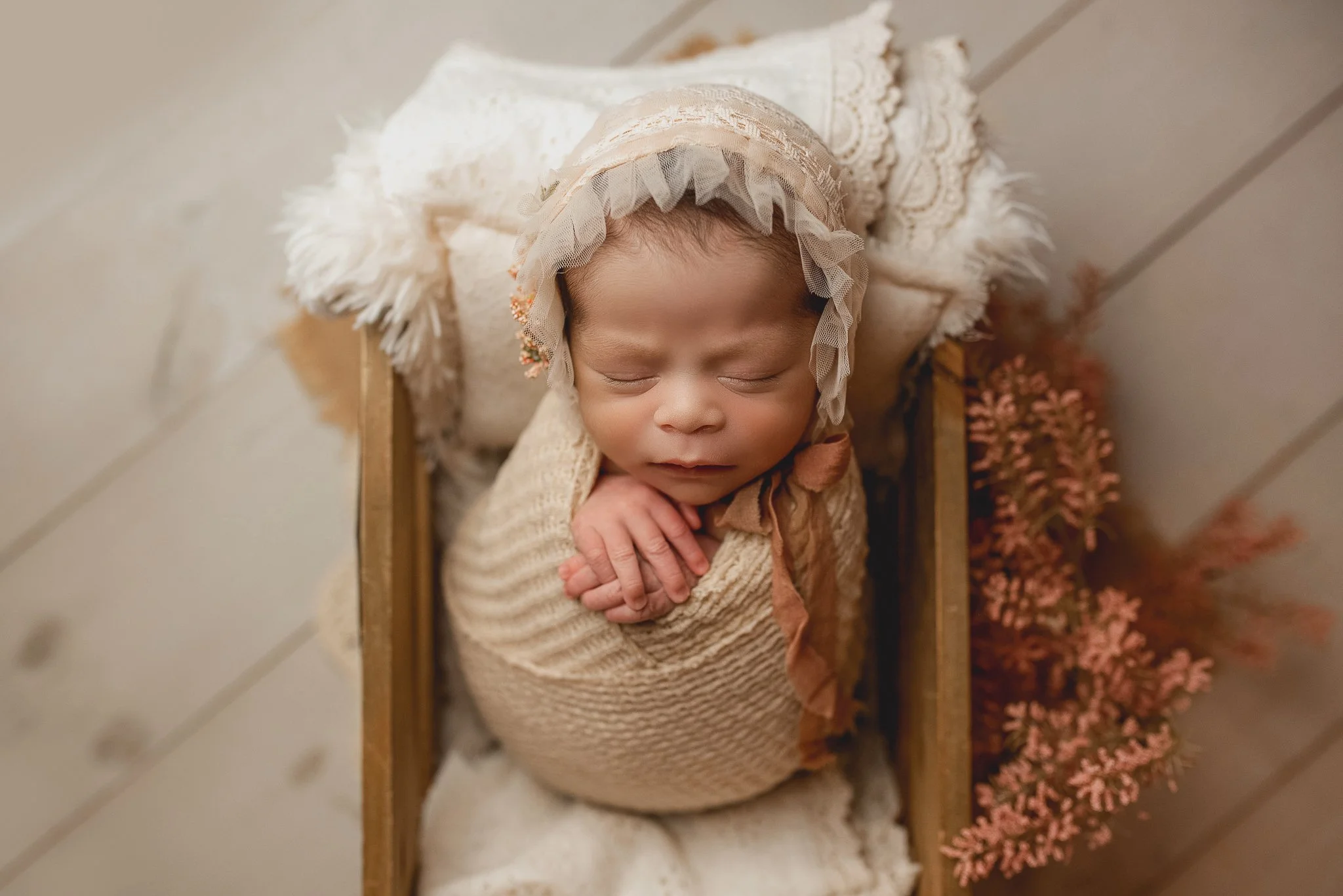 A sleeping baby wearing a vintage bonnet and cozy beige outfit, lying on a small wooden bed with a white blanket, surrounded by pink flowers.