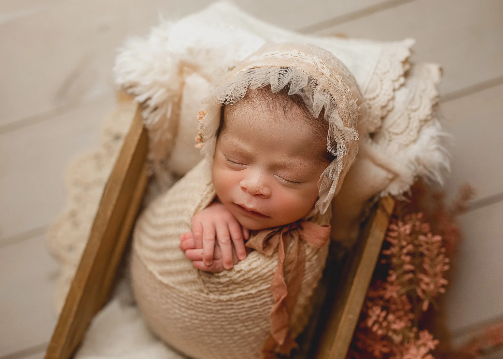 Sleeping newborn baby in a neutral bonnet posed in a wooden cradle during a newborn photography session.