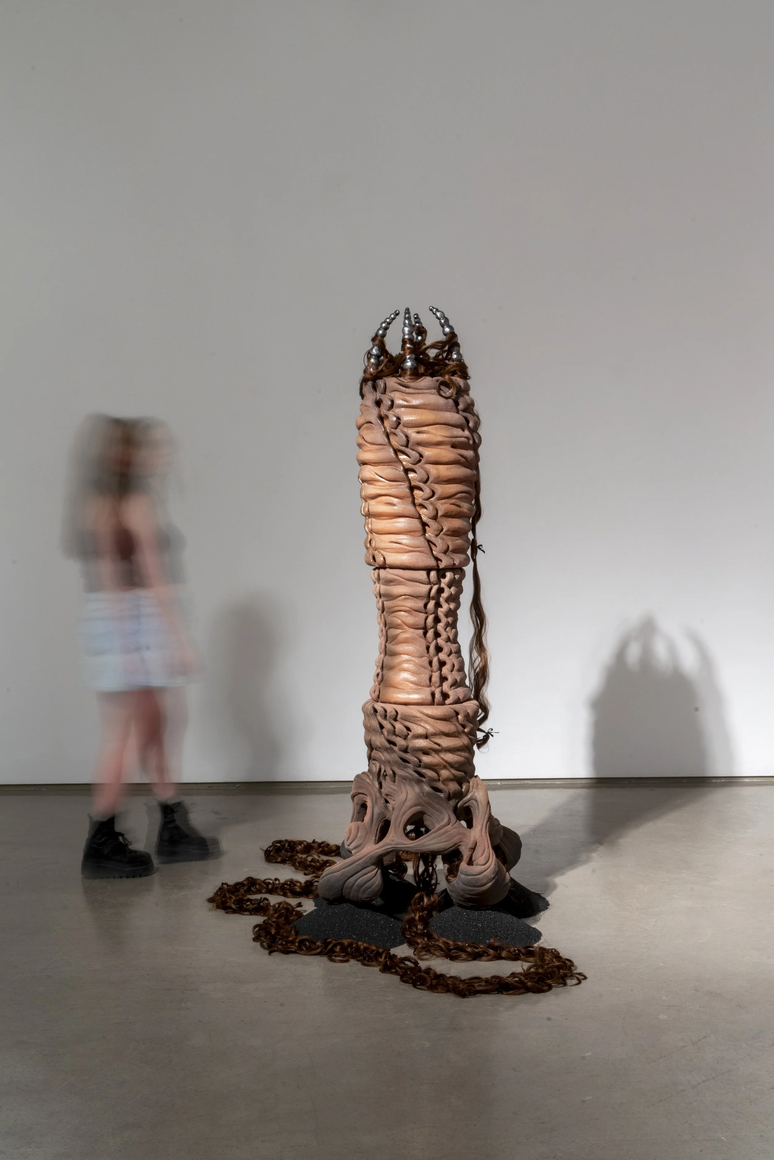 French Knitter (A Tube within a tube), 2025. Ceramic, cast aluminum, synthetic hair, sand blasting medium, polyester ribbon. 79 x 28 x 24 in.