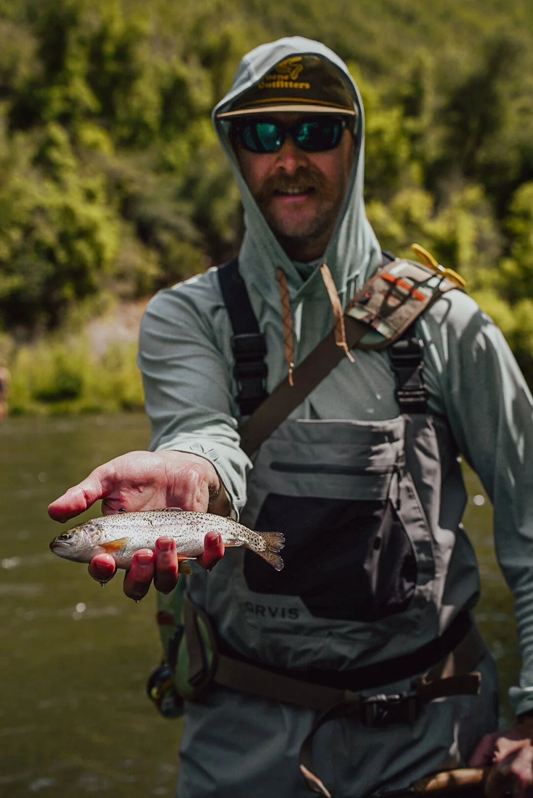 Person wearing outdoor gear and sunglasses holding a small fish.