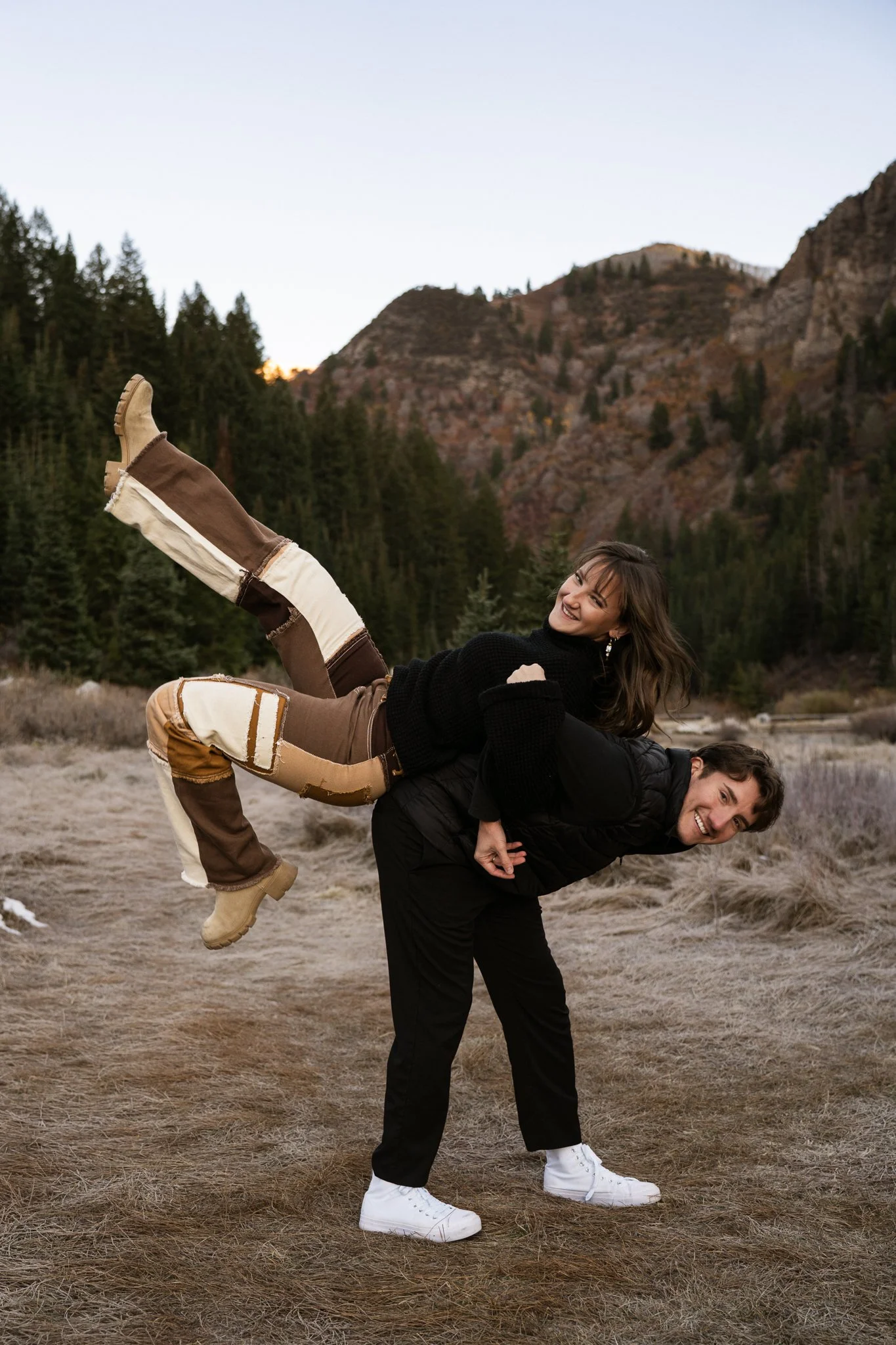 A couple enjoying an outdoor photo in a mountainous area with pine trees, where the man is bending forward with the woman sitting on his back, both smiling.
