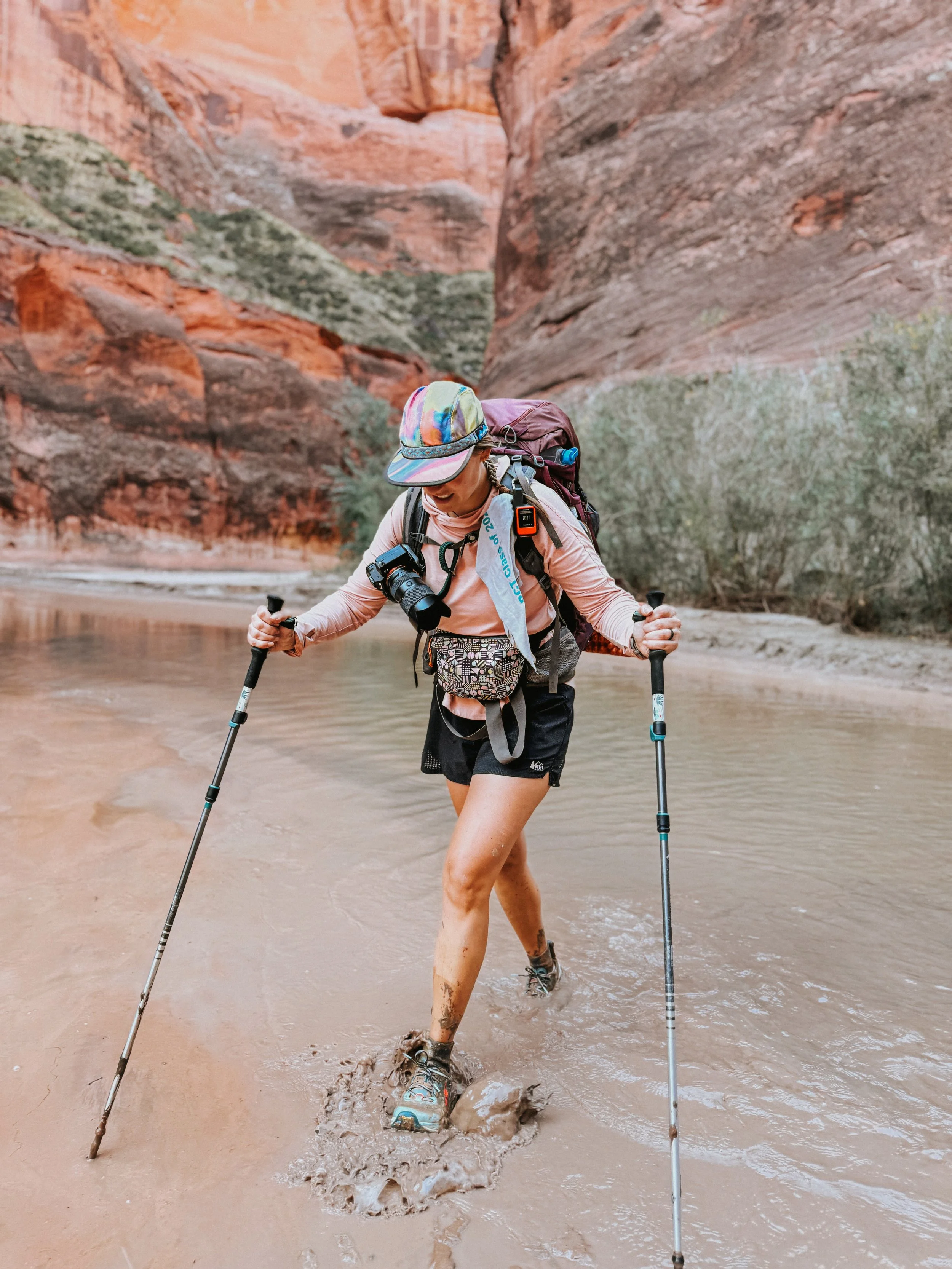 A woman hiking through a muddy creek surrounded by sandstone cliffs, wearing a colorful hat, sunglasses, a backpack, and using trekking poles.