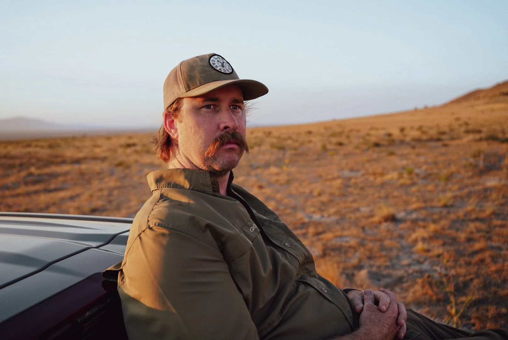 A man with a mustache and brown hair, wearing a tan cap and an olive-green jacket, sitting outdoors in a dry, hilly landscape during sunset.