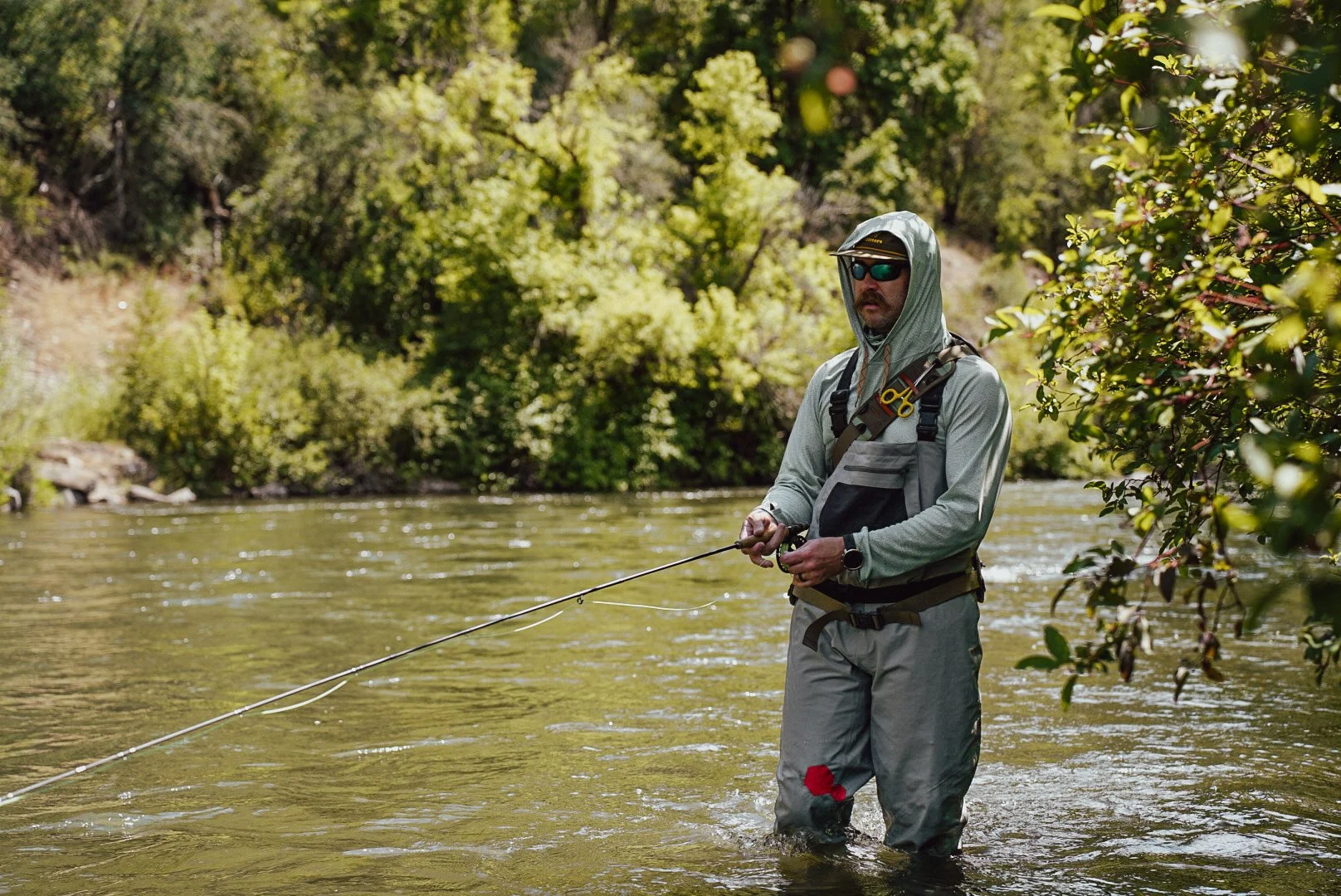 Man fishing in a river, dressed in outdoor gear, sunglasses, and a hoodie, amidst lush green trees.