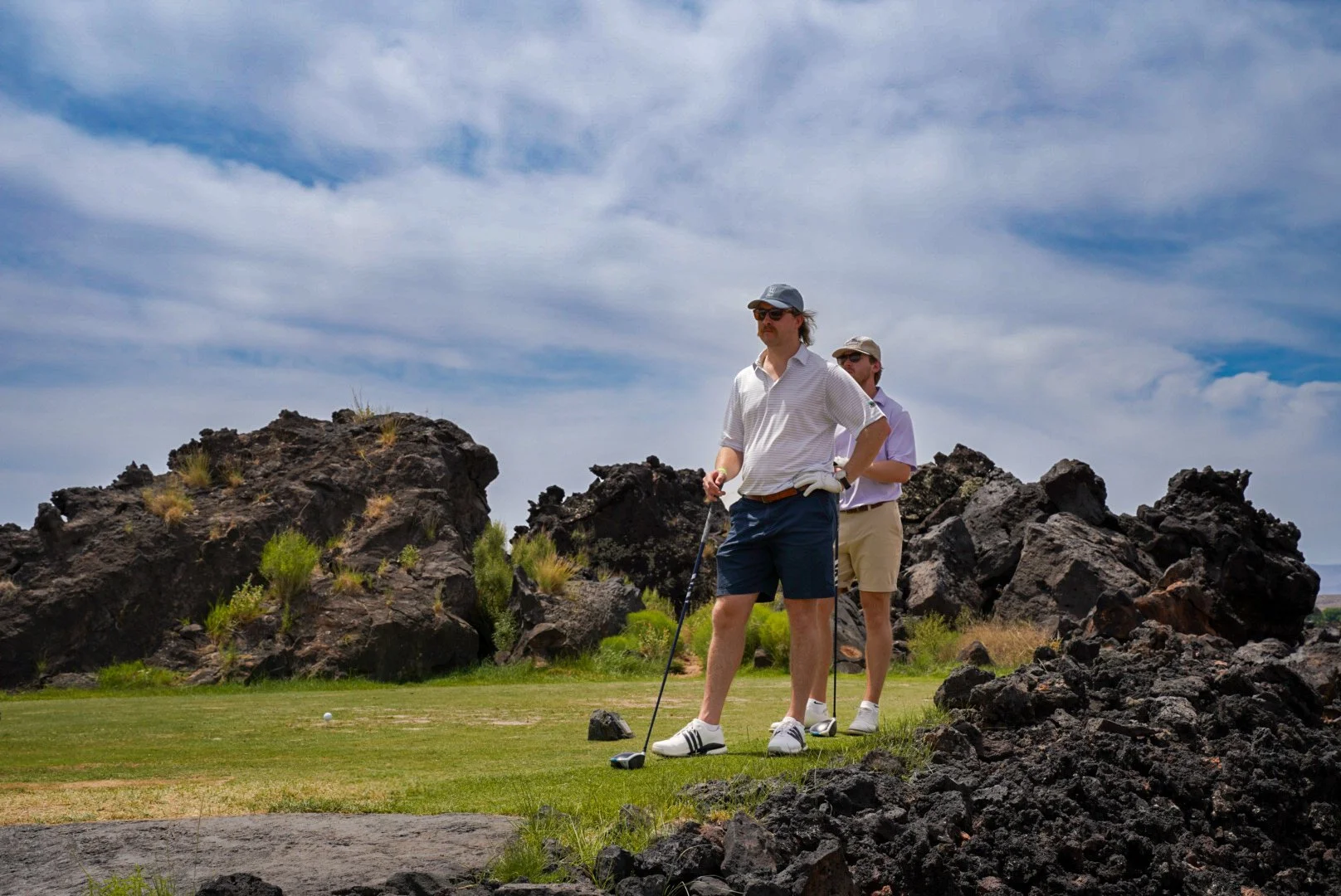 Two men playing golf on a course with large rocks and grass, under a partly cloudy sky.