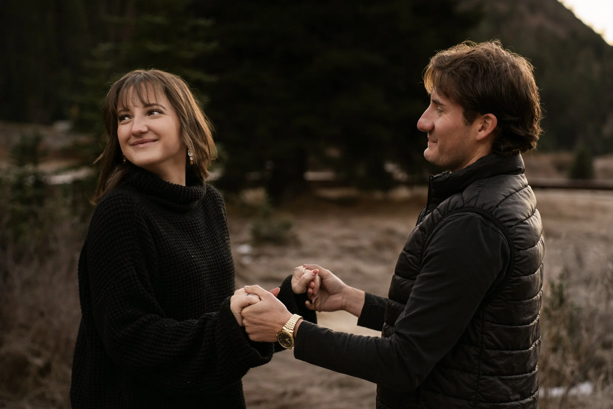 A young woman with shoulder-length brown hair and a black sweater holding hands with a young man wearing a black puffer vest outdoors, smiling at each other against a backdrop of trees and a dirt path.