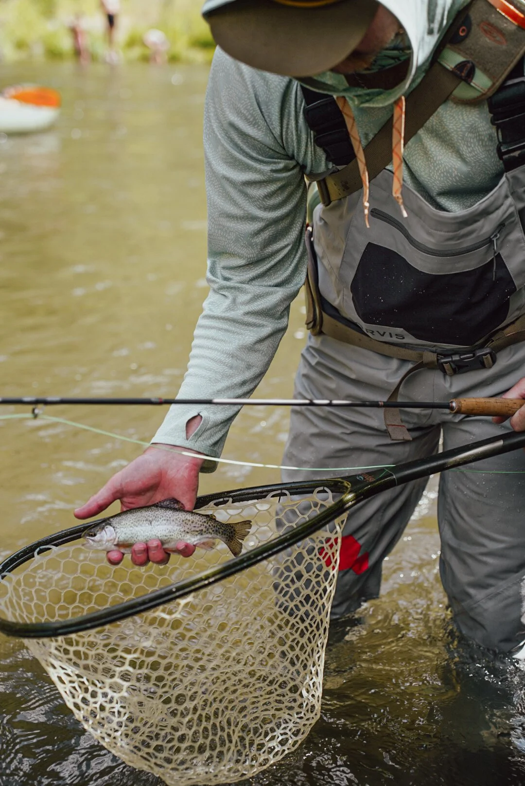 Person holding a small fish in a fishing net while standing in water, with fishing gear and outdoor attire.