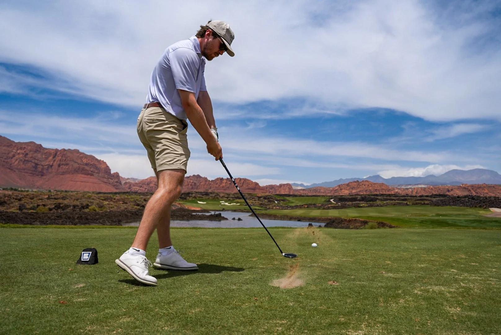 Man in athletic clothing playing golf on a course with a landscape of red mountains and blue skies in the background.