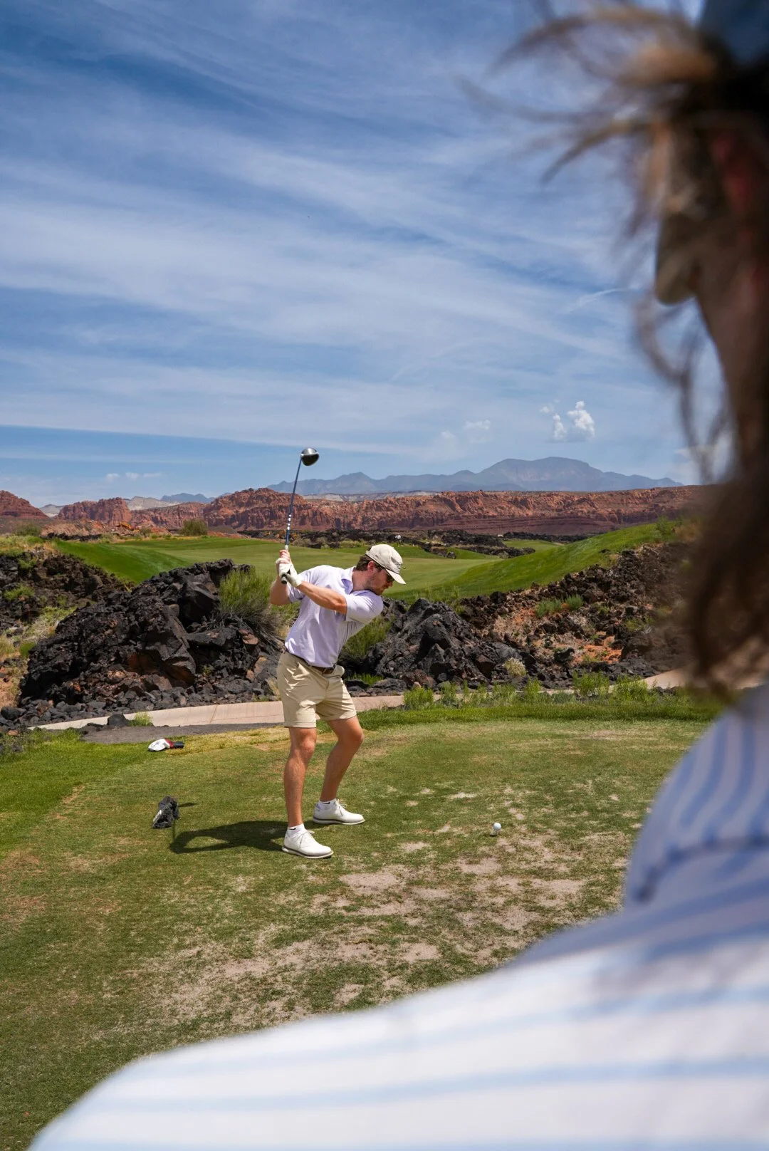 A man in a white shirt and beige shorts preparing to hit a golf ball on a golf course with mountainous desert landscape in the background.
